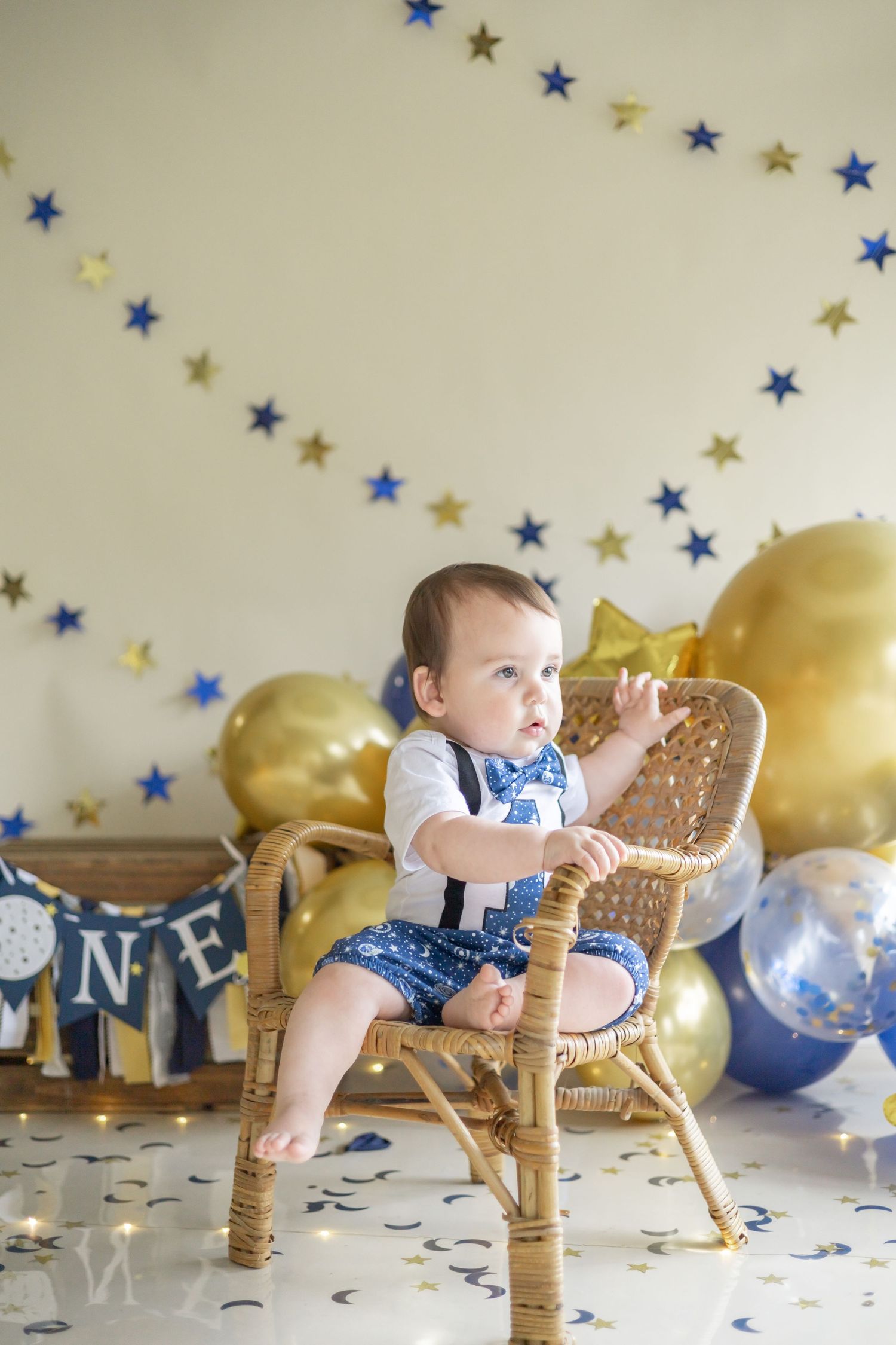 Baby in blue outfit sitting in rattan chair with gold balloons and blue star decorations in background for milestone photo.