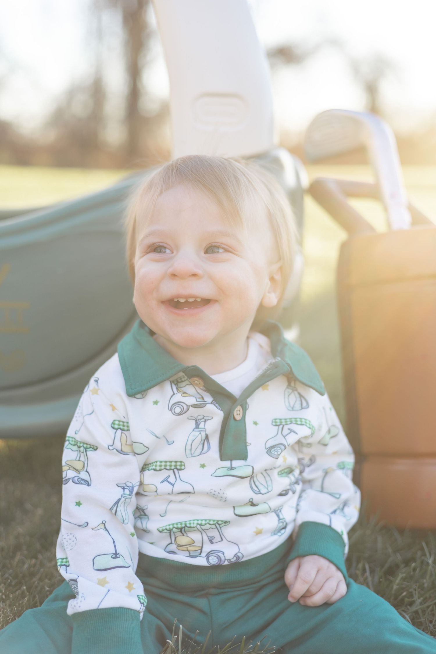 A small child wearing a white dinosaur print shirt and green pants sits on grass in golden sunlight.