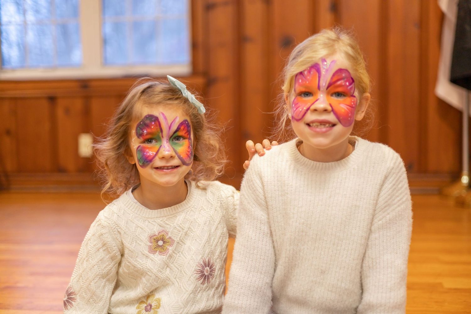 Face-painted children in white sweaters at a festive holiday gathering in a wood-paneled room.