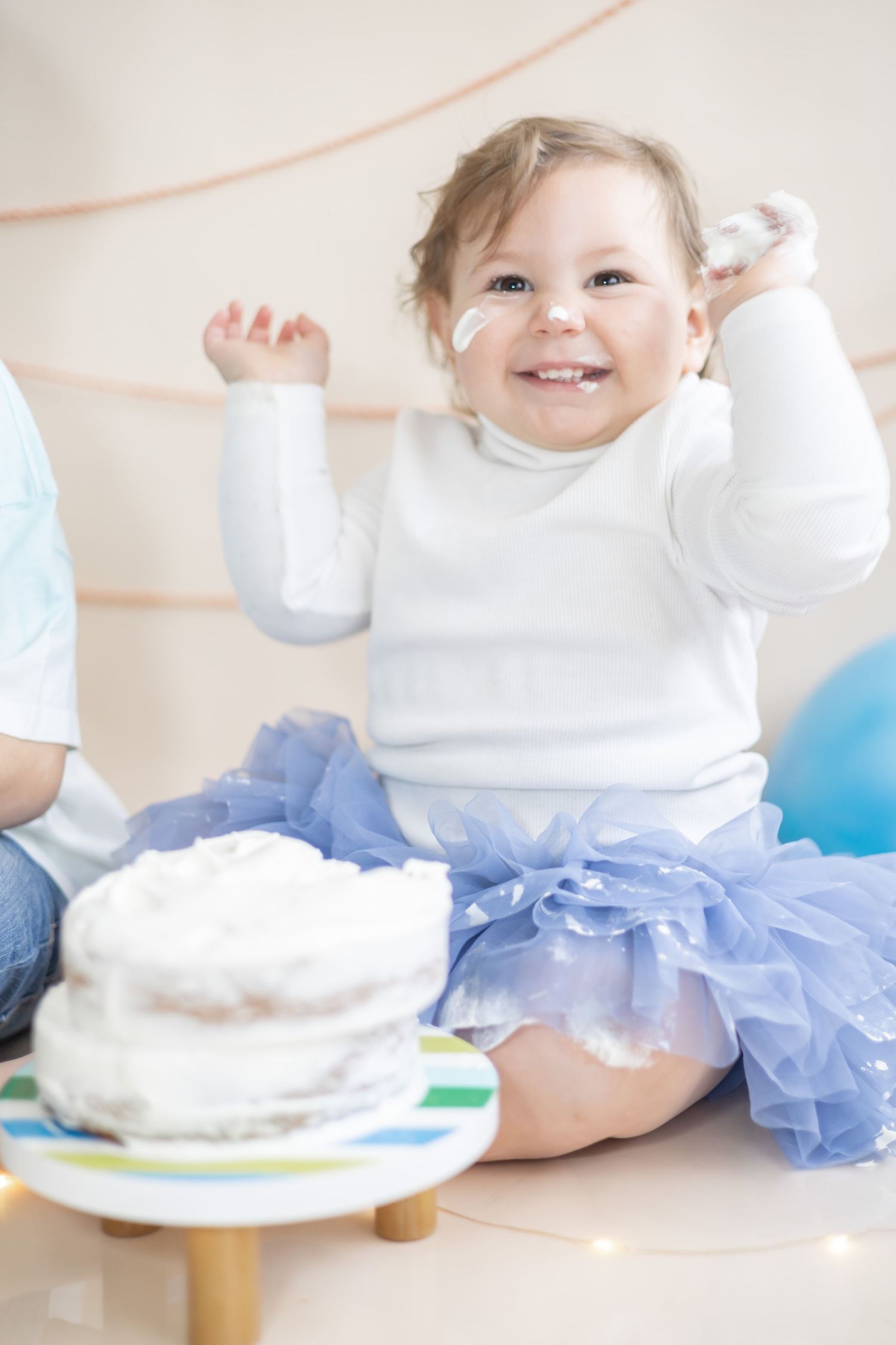 Baby in white sweater and blue tutu sitting on floor with smash cake during first birthday photoshoot.