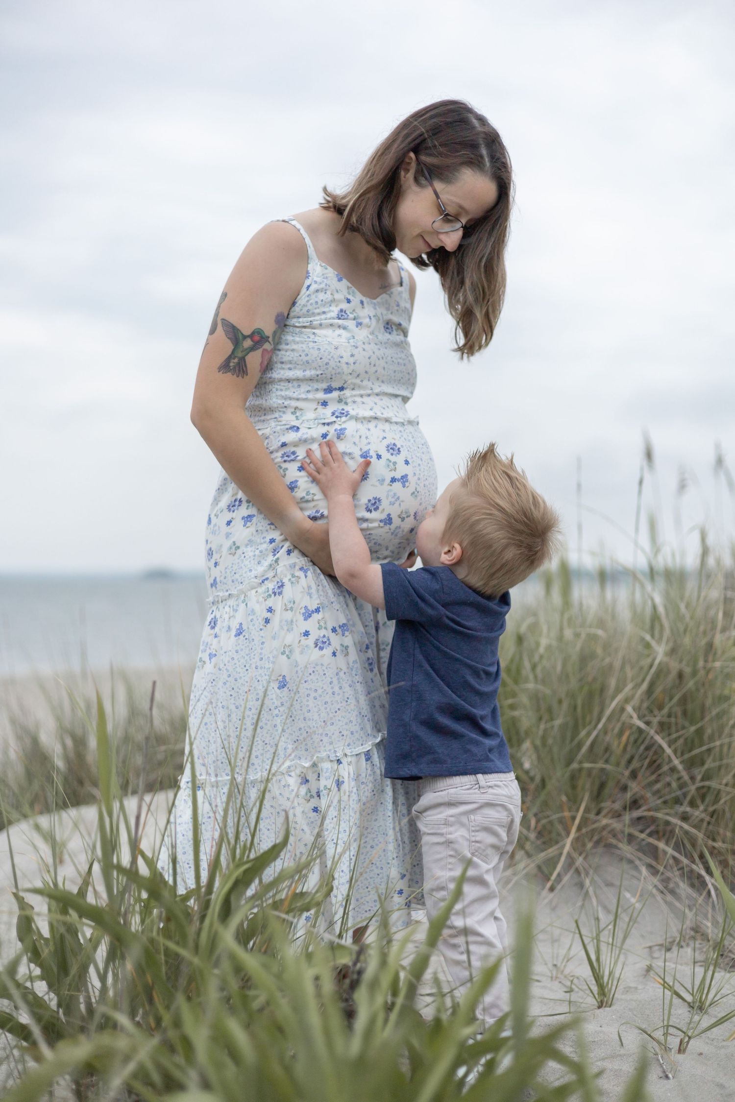 A tender moment between parent and child standing among beach grass with the ocean visible in the background.