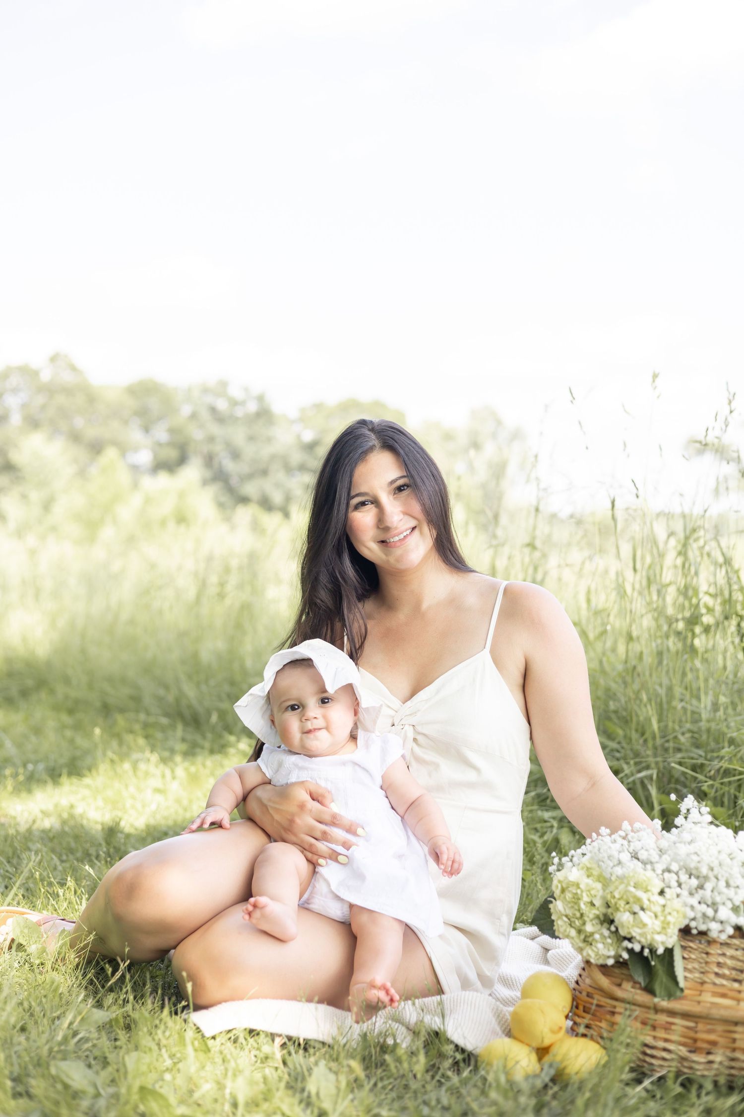 A mother and baby sitting in a sunny meadow with a wicker basket and yellow rain boots beside them.