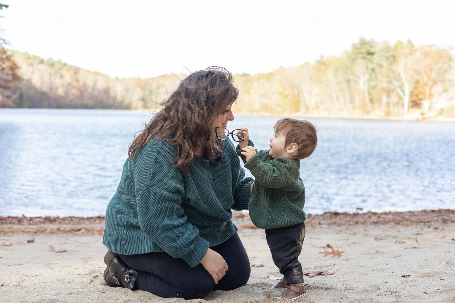 A mother and child sit together on a sandy beach near a lake on an autumn day wearing matching green sweaters.
