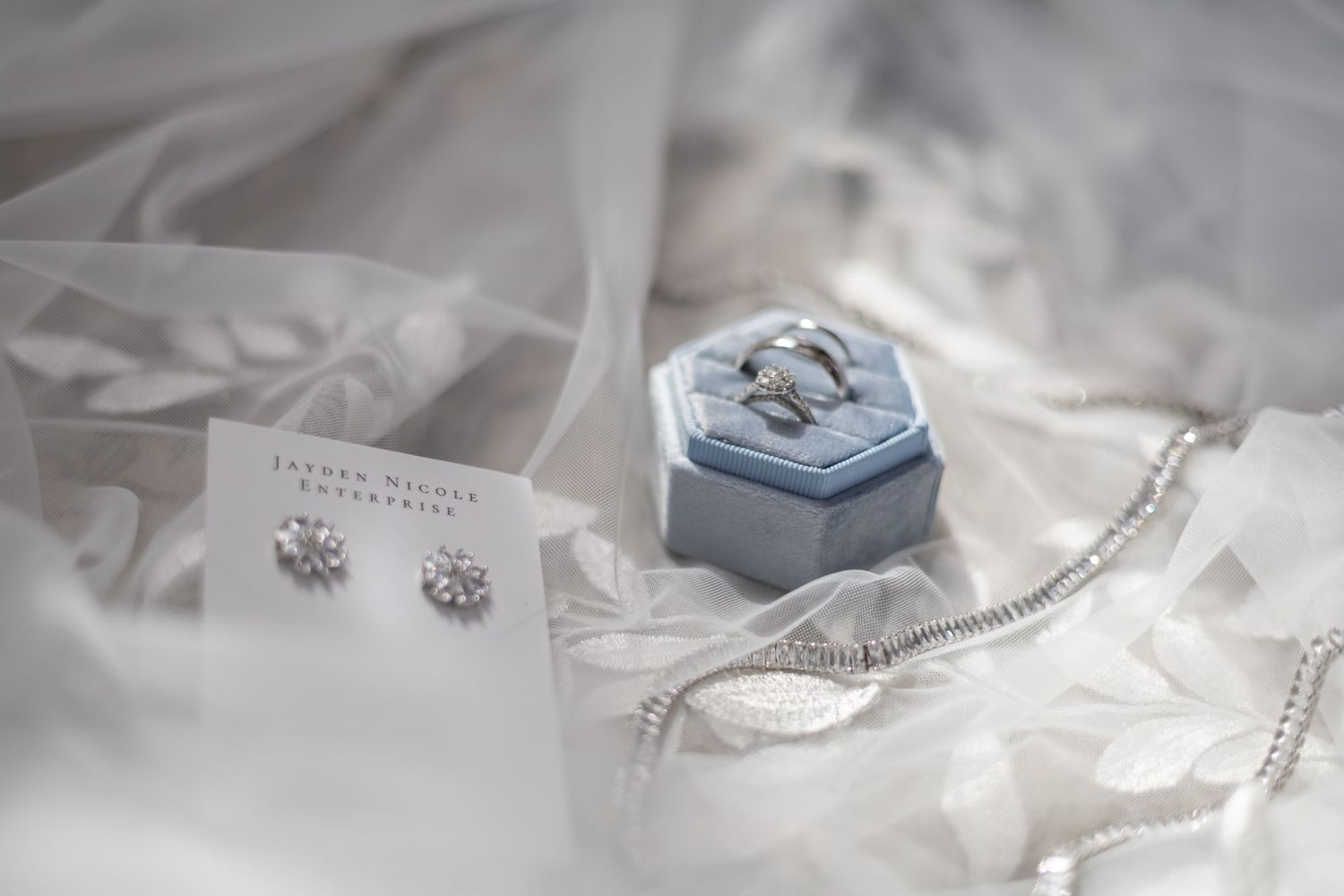 A delicate arrangement of wedding rings, diamond earrings, and pearl jewelry on soft white fabric with ethereal lighting.