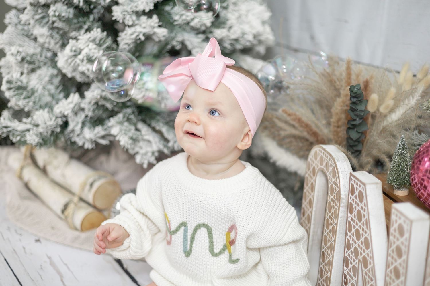 Small child wearing white sweater with 'one' text and pink bow headband sitting by frosted Christmas tree.