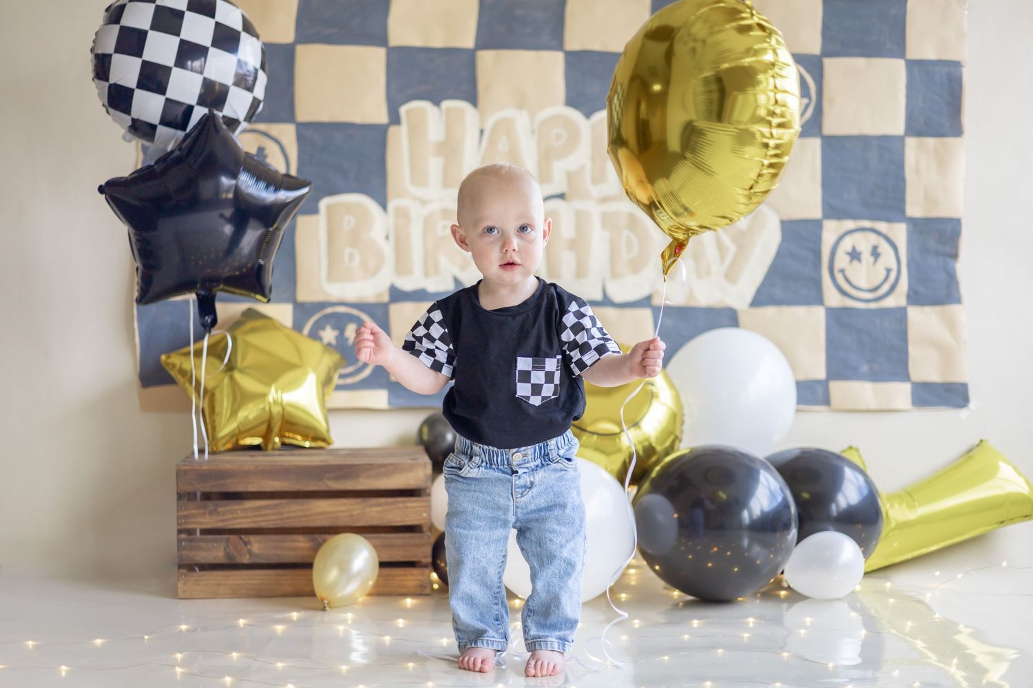 Baby's first birthday celebration setup with black and gold balloons, checkered backdrop, and wooden crates.