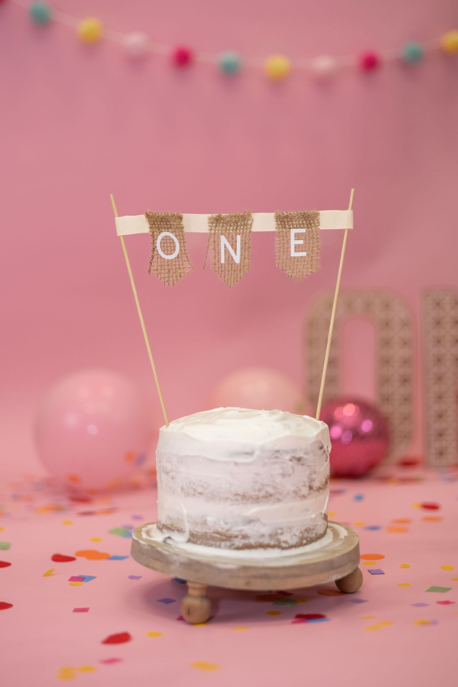 A small white birthday cake with 'ONE' banner topper sits on wooden stand against pink backdrop with colorful pom pom garland.