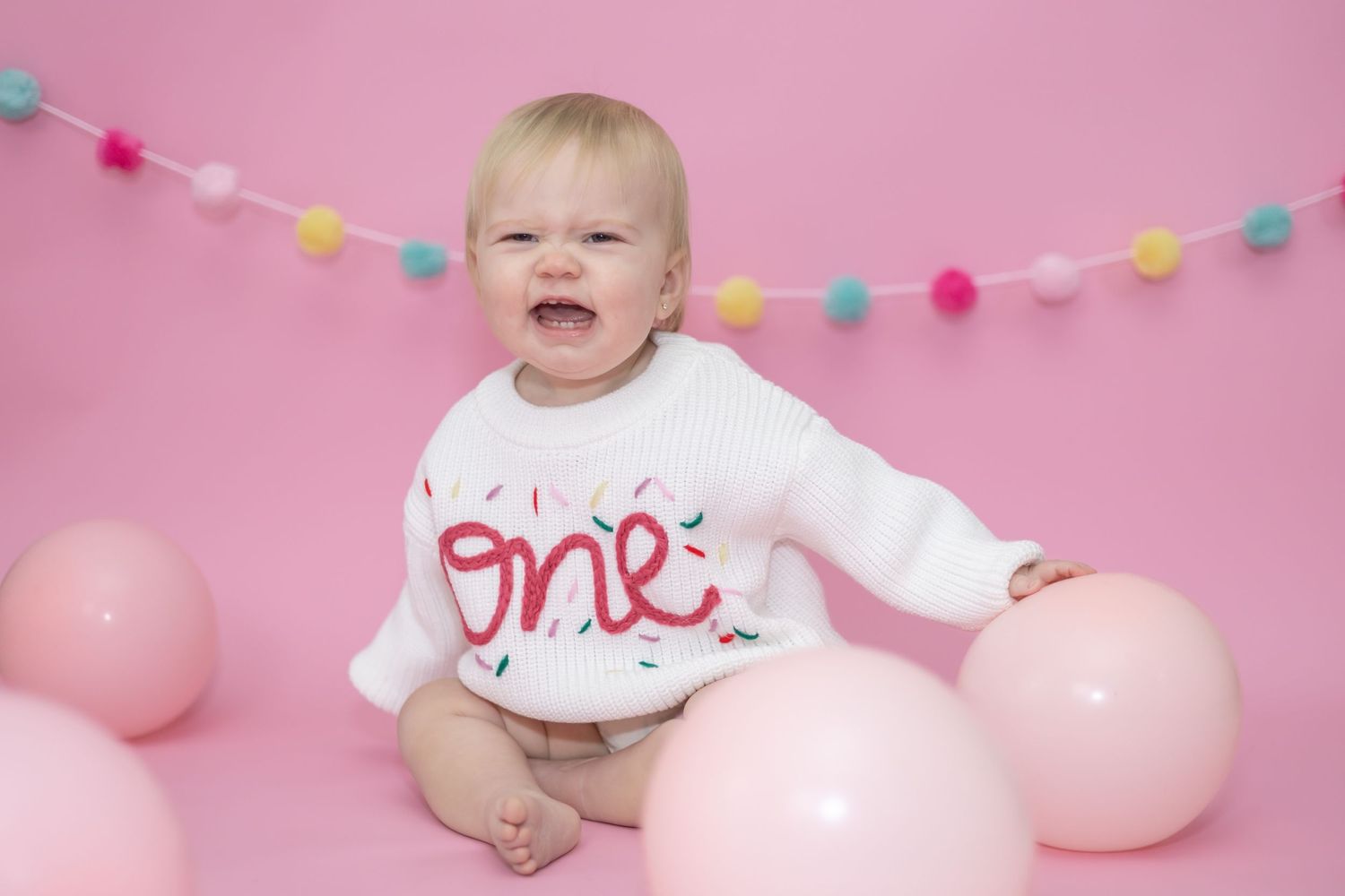 Small baby wearing white onesie lying on pink backdrop with colorful party decorations and balloons.