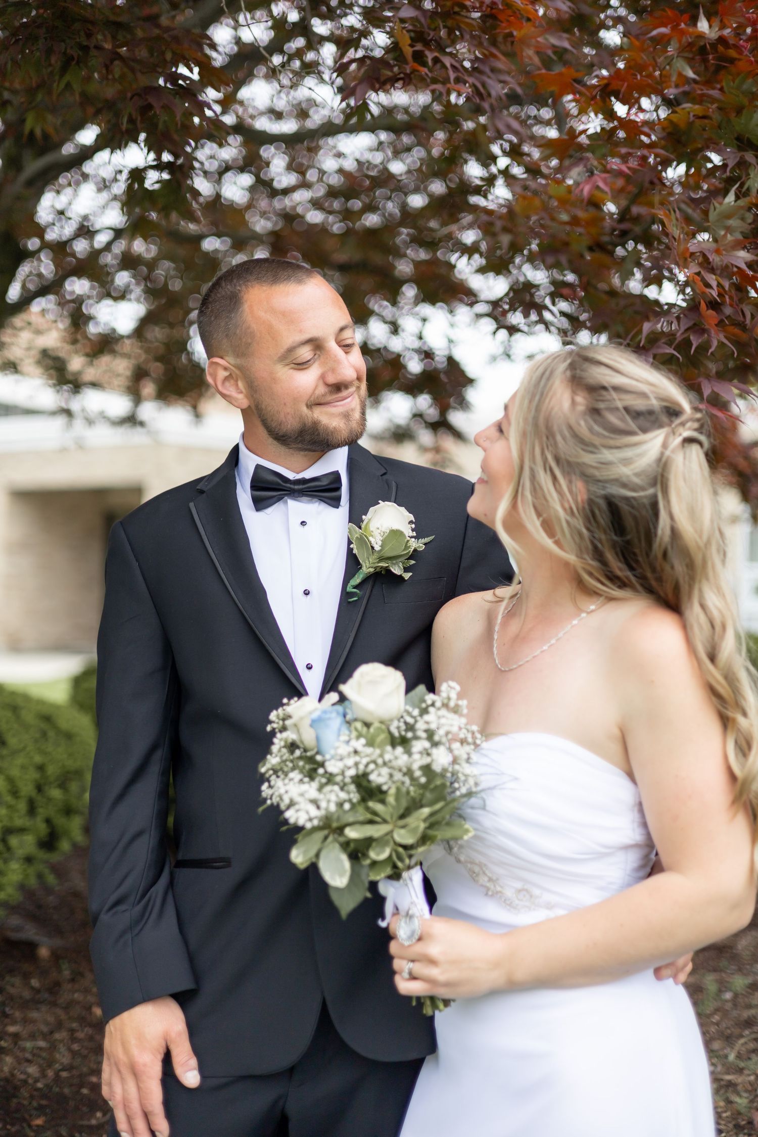 A couple shares a loving moment at their wedding beneath autumn trees, with the bride holding a delicate white bouquet.