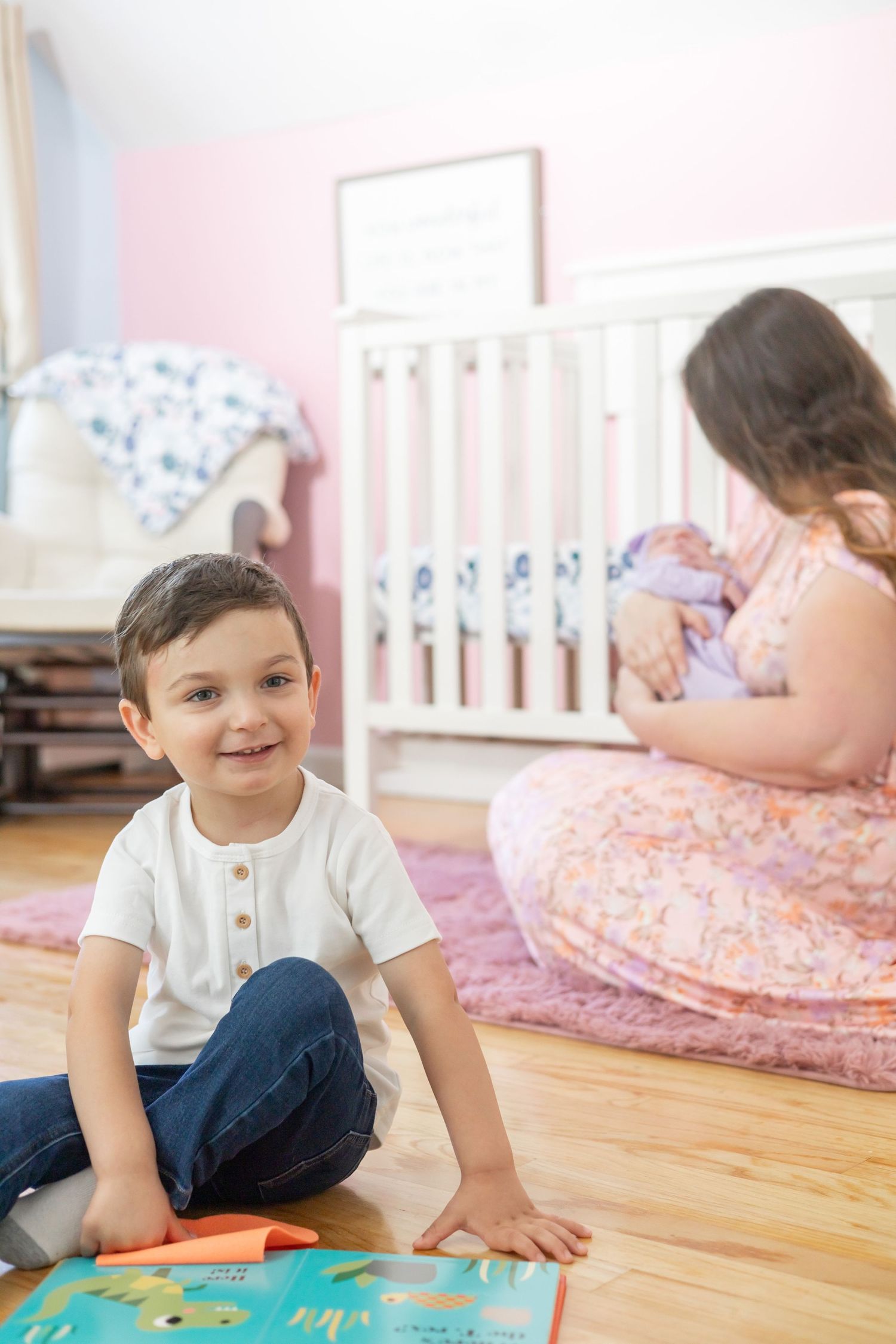 Children in nursery room with pink walls, white crib, and educational mat on hardwood floor.