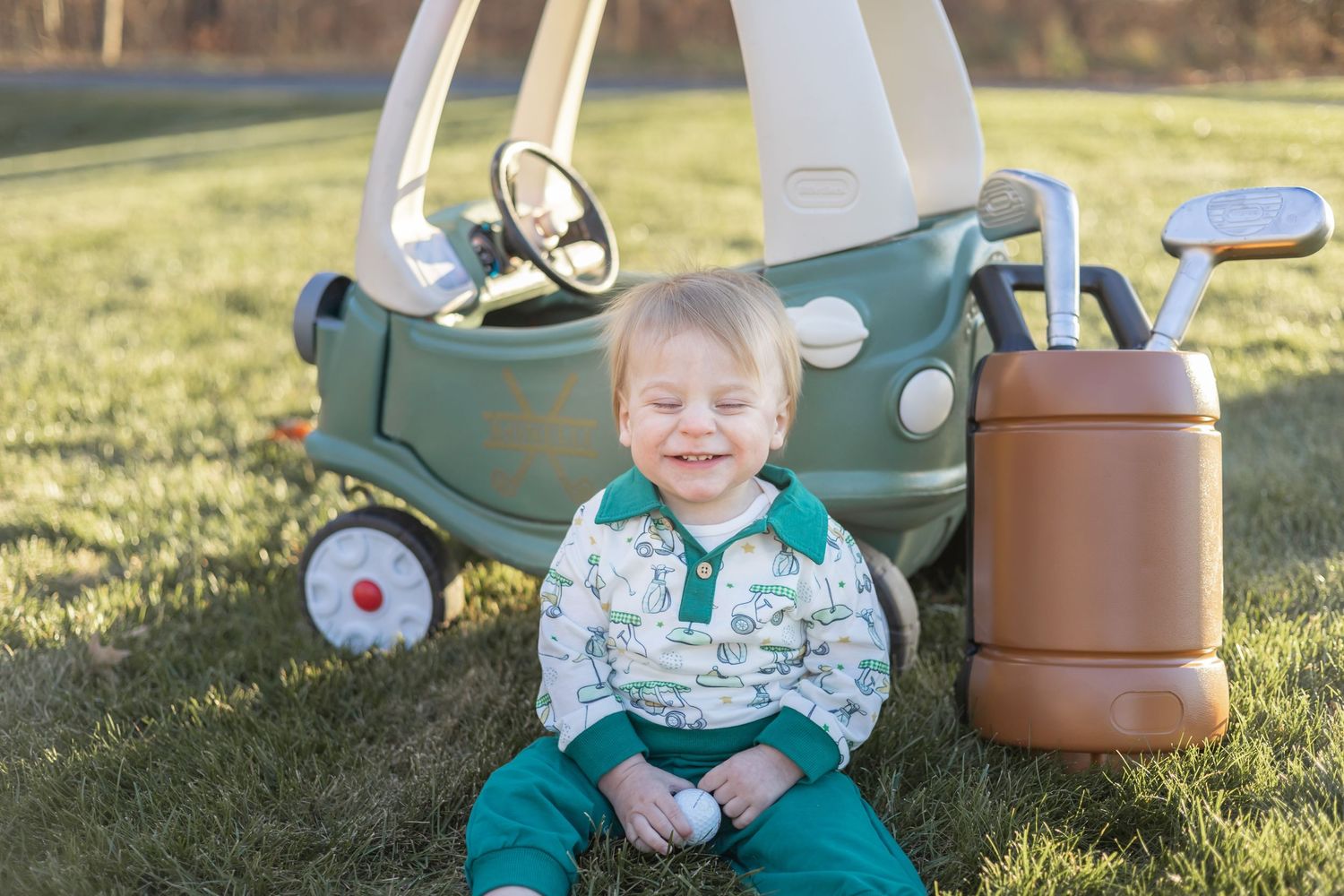 A toddler plays with a vintage-style green toy car while sitting in grass on a sunny afternoon.