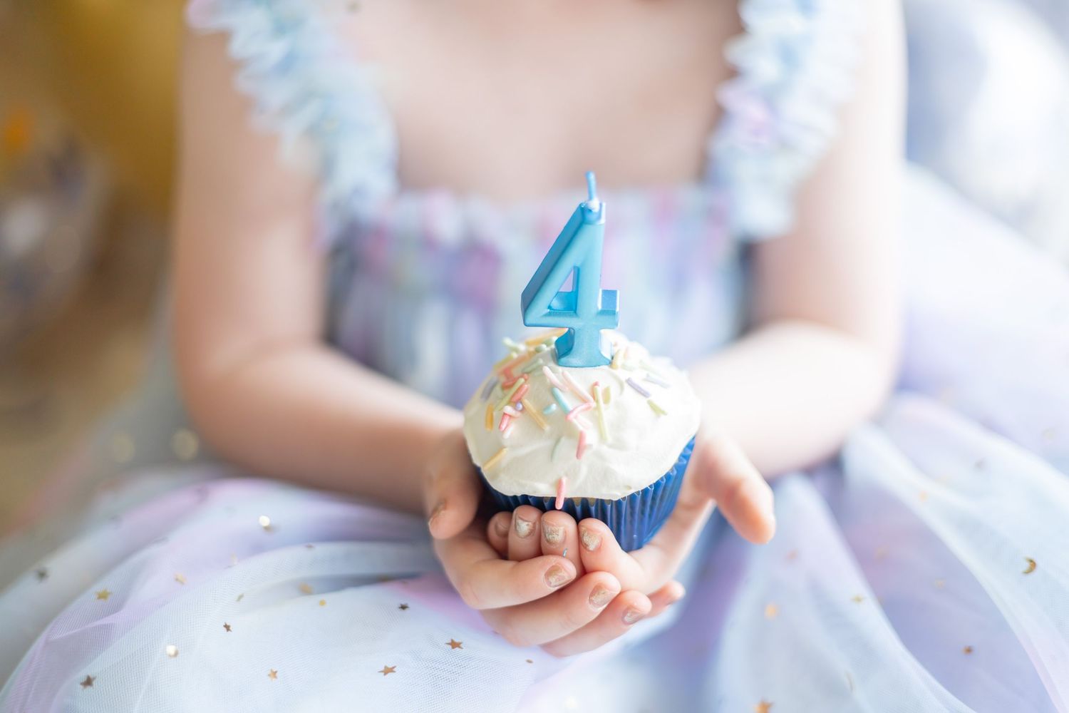 A close-up shot of hands holding a blue cupcake decorated with a white number 4 candle and sprinkles.