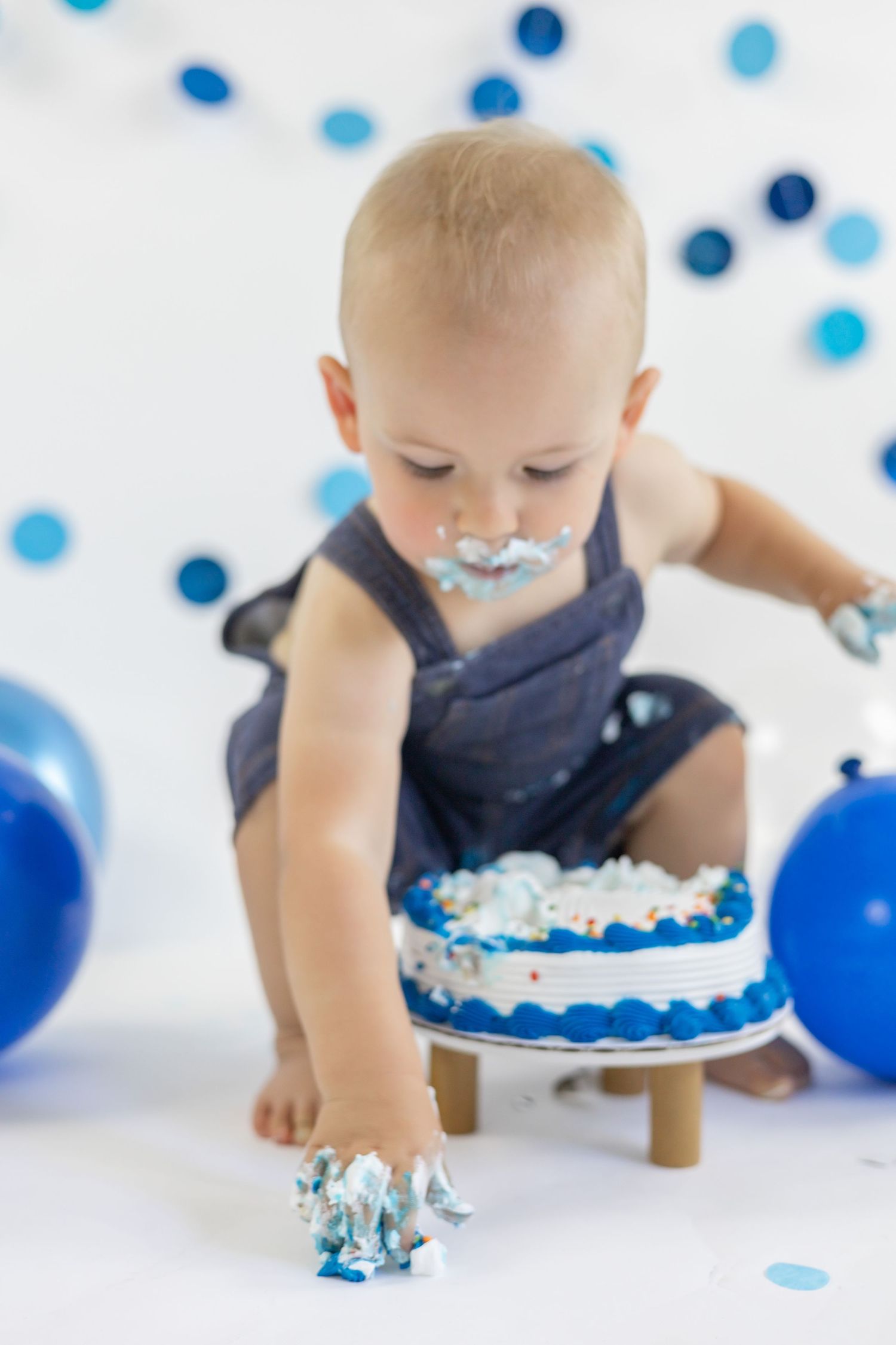 Baby in blue overalls smashes into birthday cake during cake smash photoshoot with blue balloon decorations.