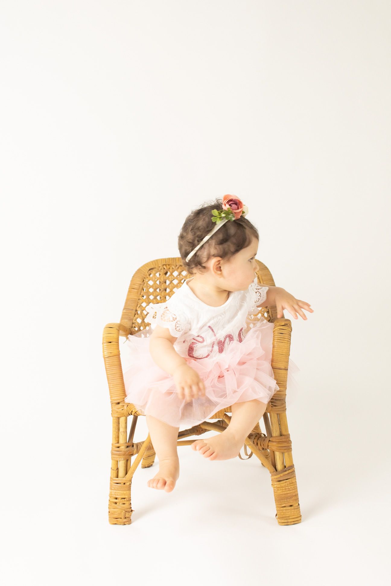 Child in pink tulle dress poses playfully on wooden chair against white backdrop.