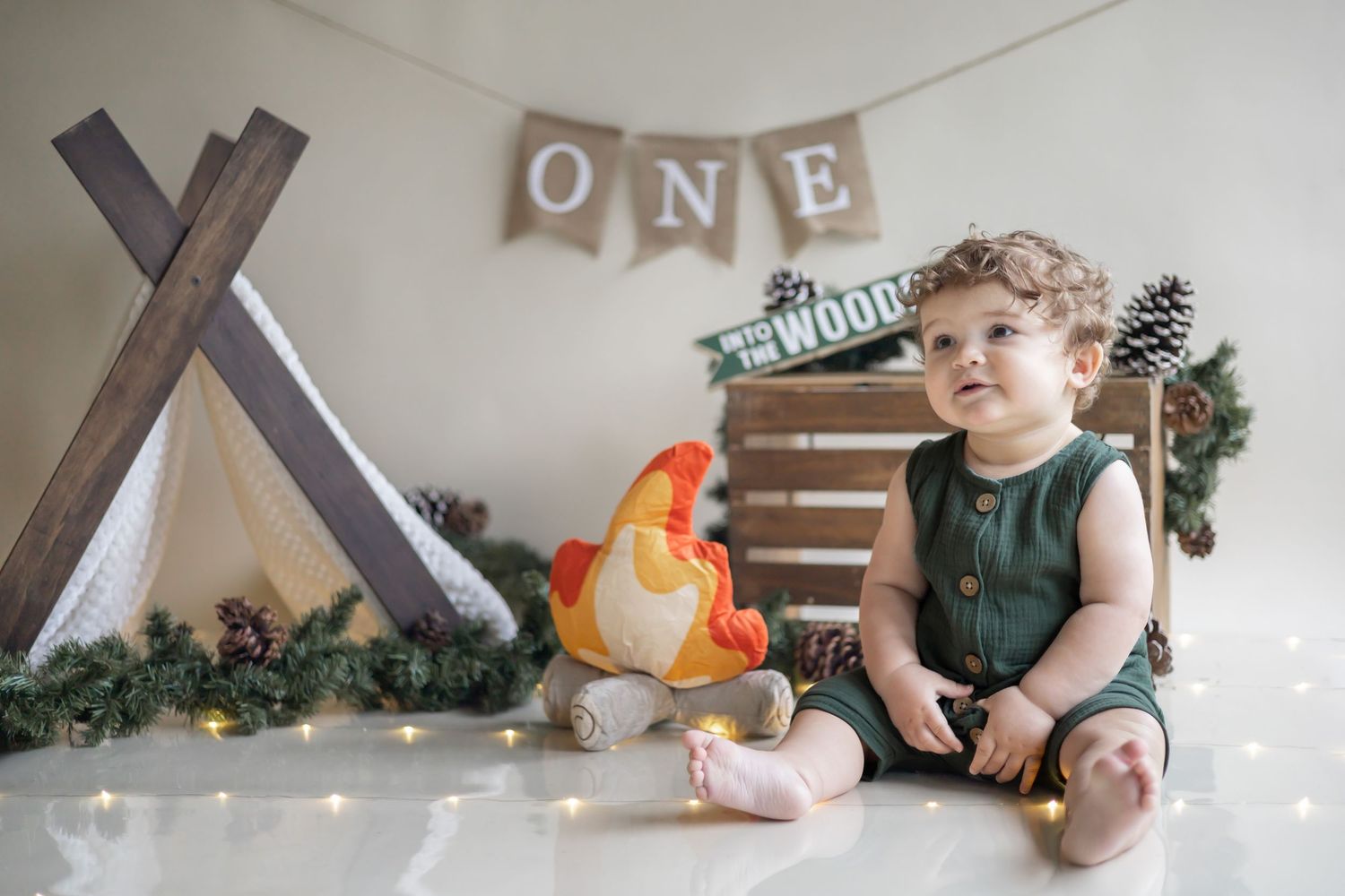 Baby in green overalls sits near wooden teepee frame with orange plush campfire and 'ONE' banner decoration.