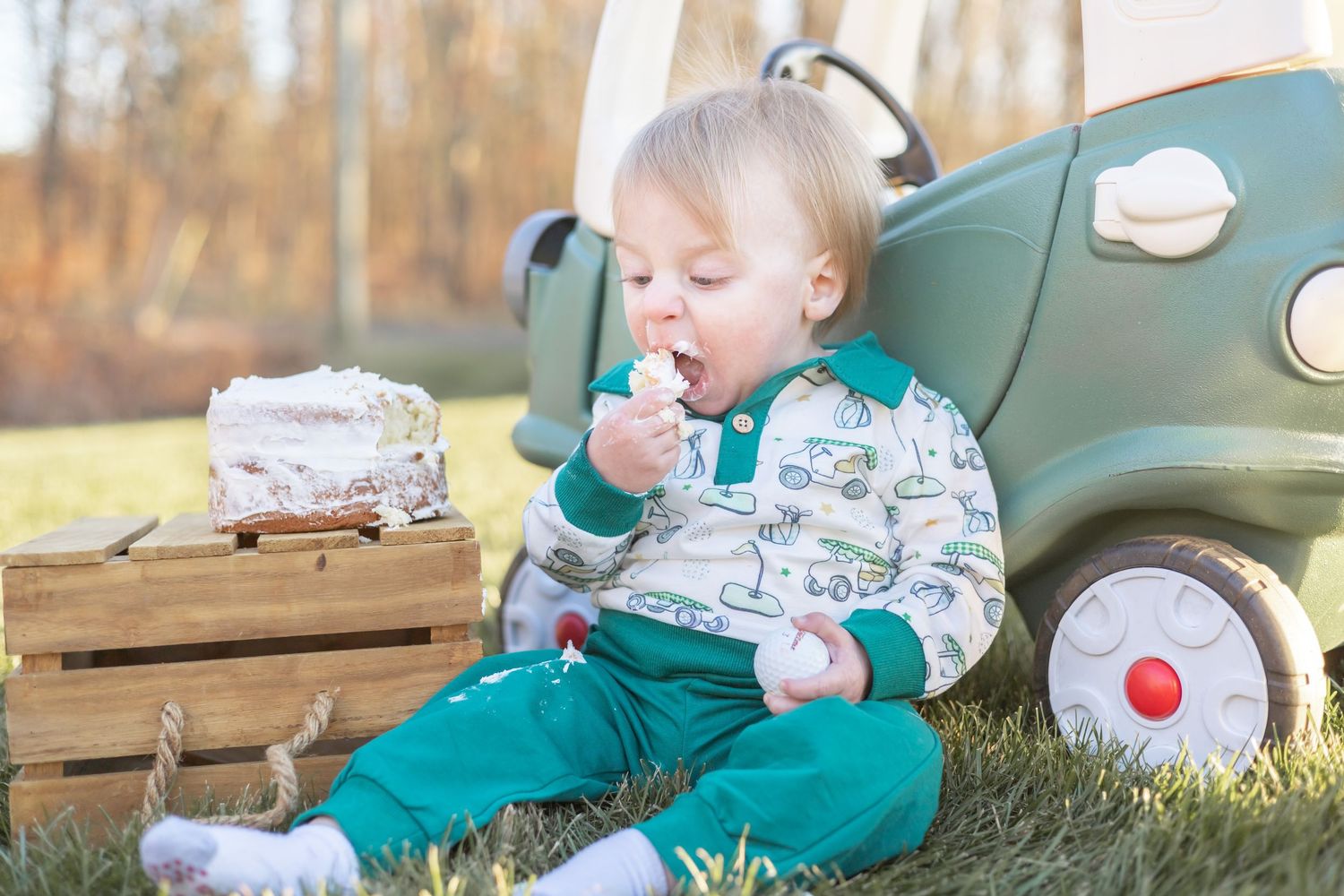 Baby in green shirt sits outdoors on blanket with toys during sunny autumn day in countryside setting.