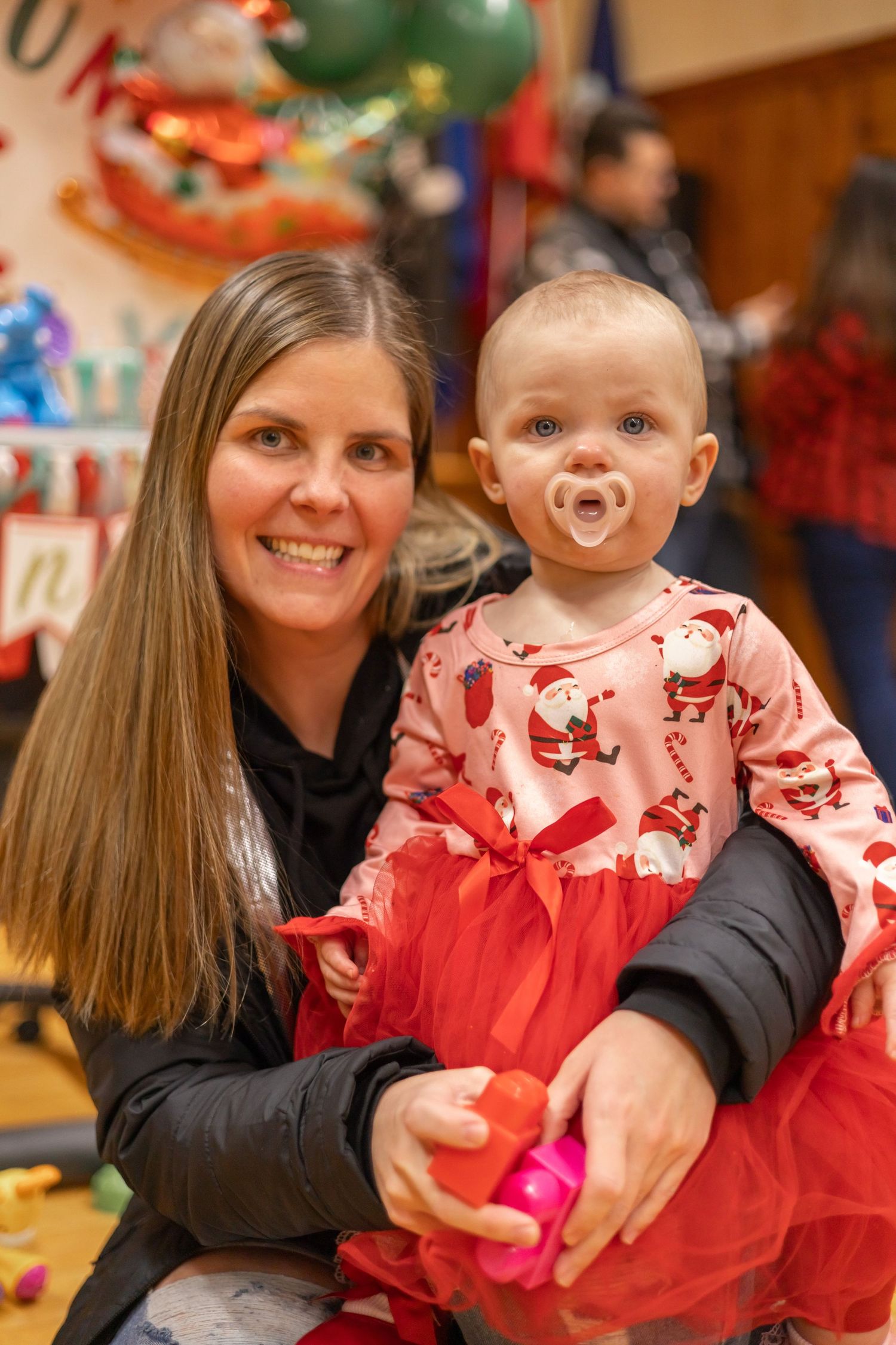 A baby wearing a festive red and white holiday dress with a pacifier sits on an adult's lap at a Christmas celebration.