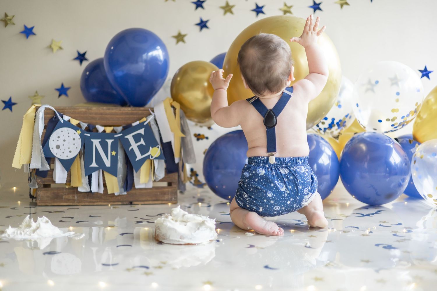 A baby in blue overalls sits on the floor surrounded by blue and gold balloons and decorations for a first birthday celebration.