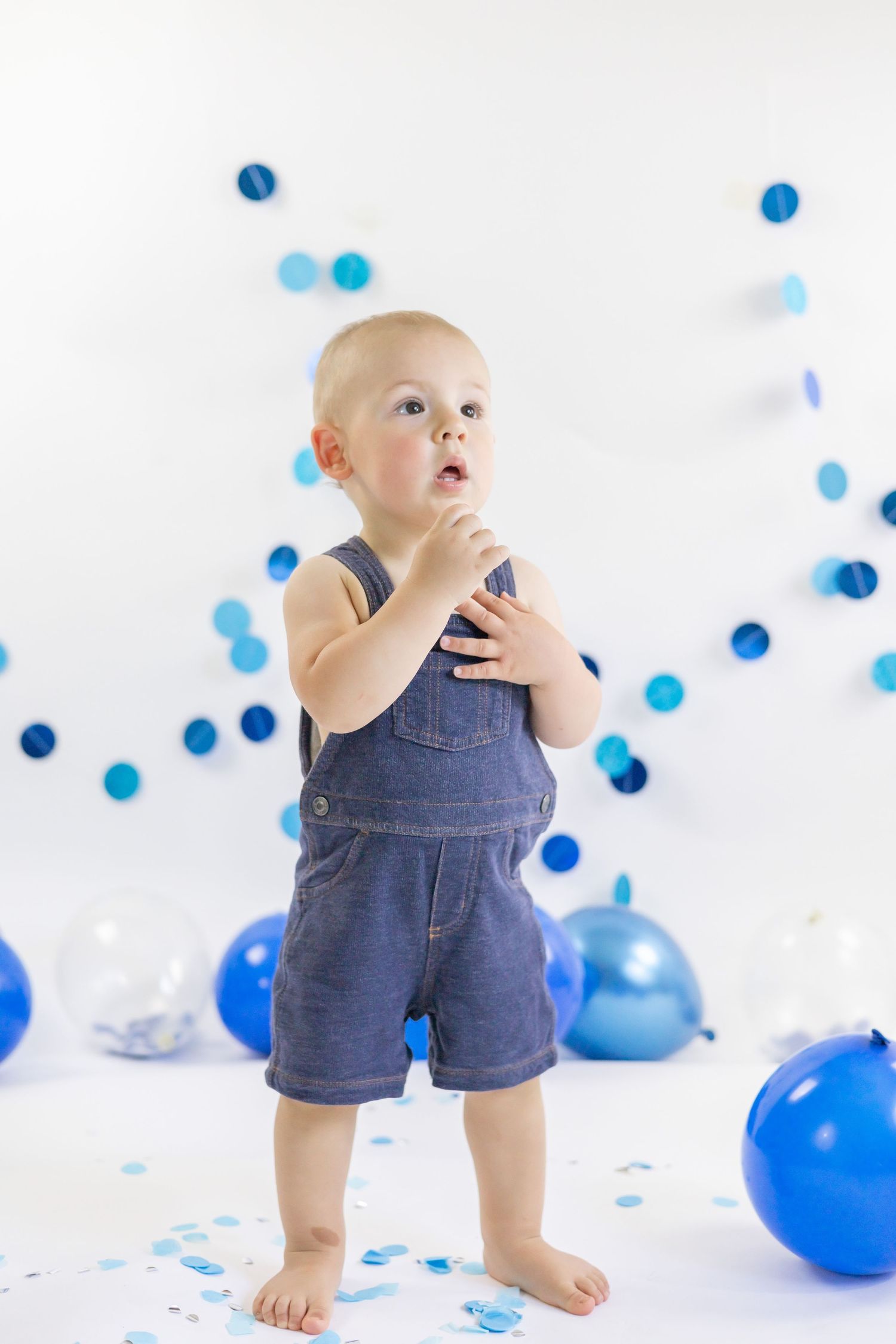Adorable baby in denim overalls plays with scattered blue balloons against a white background.