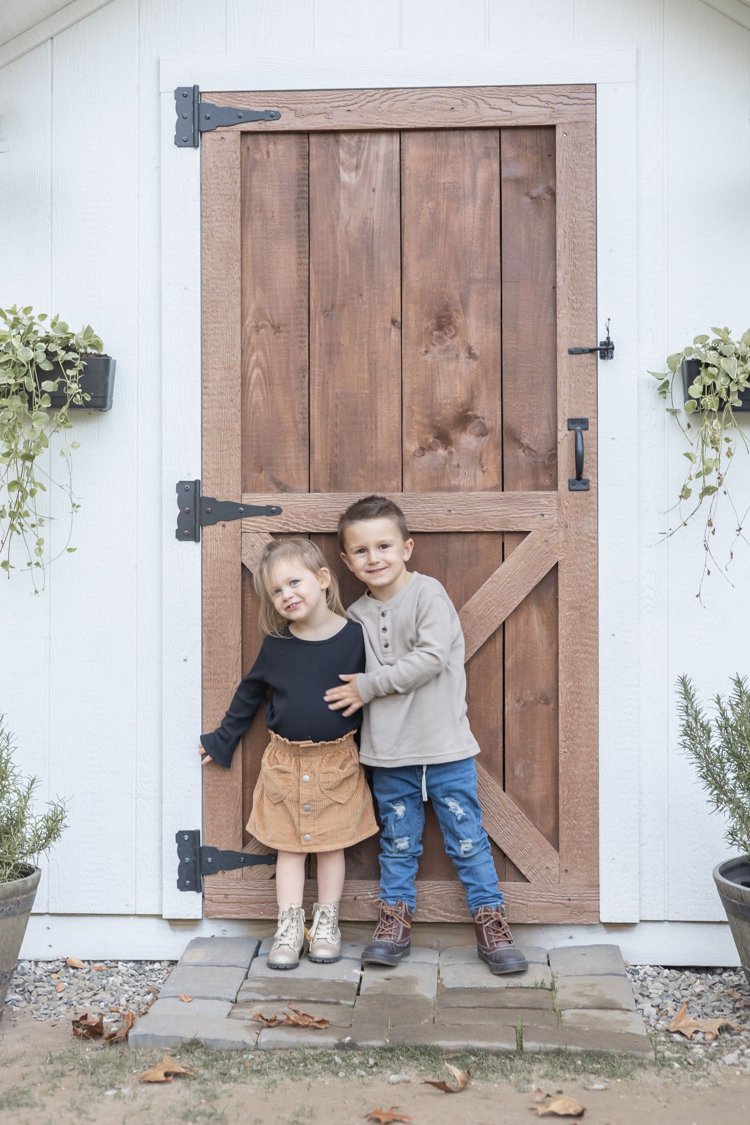 Children sitting and playing by a rustic wooden barn door against a white wall with hanging plants.