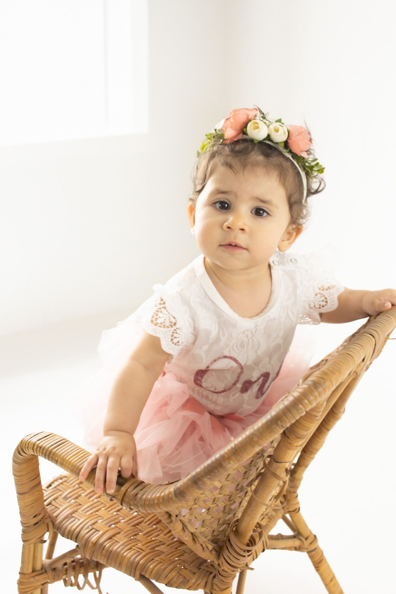 Child in pink tutu dress and floral headband sits on woven rattan chair against white background.