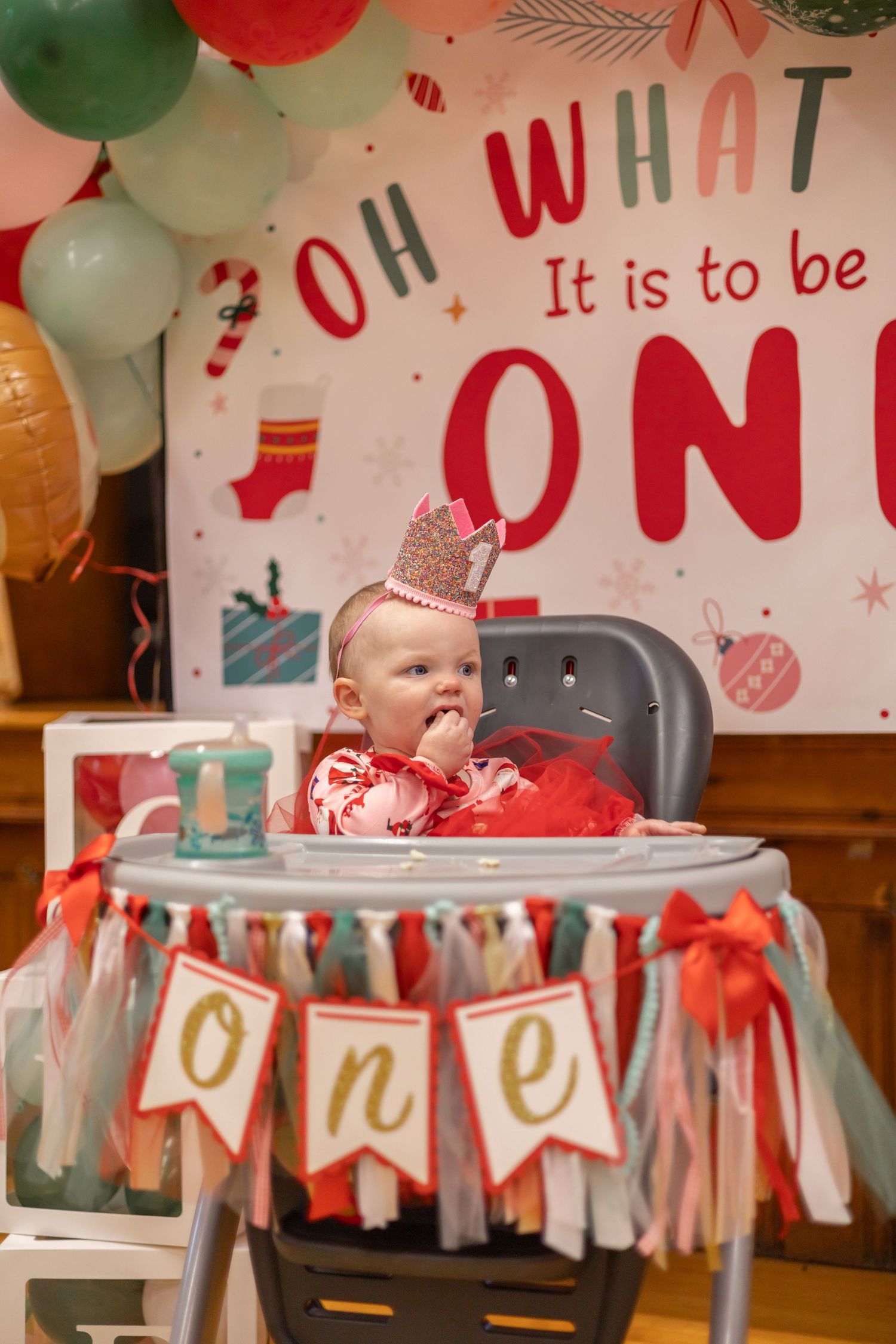 A baby sits in a high chair decorated with a festive banner and balloons for a first birthday celebration.