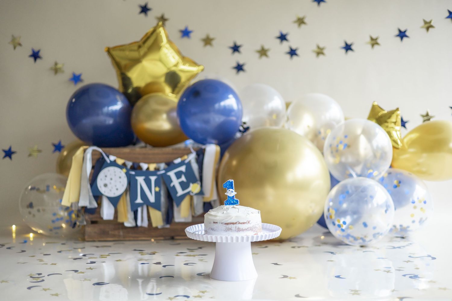 First birthday party decoration setup with blue and gold balloons, silver confetti, and a white cake stand on marble surface.