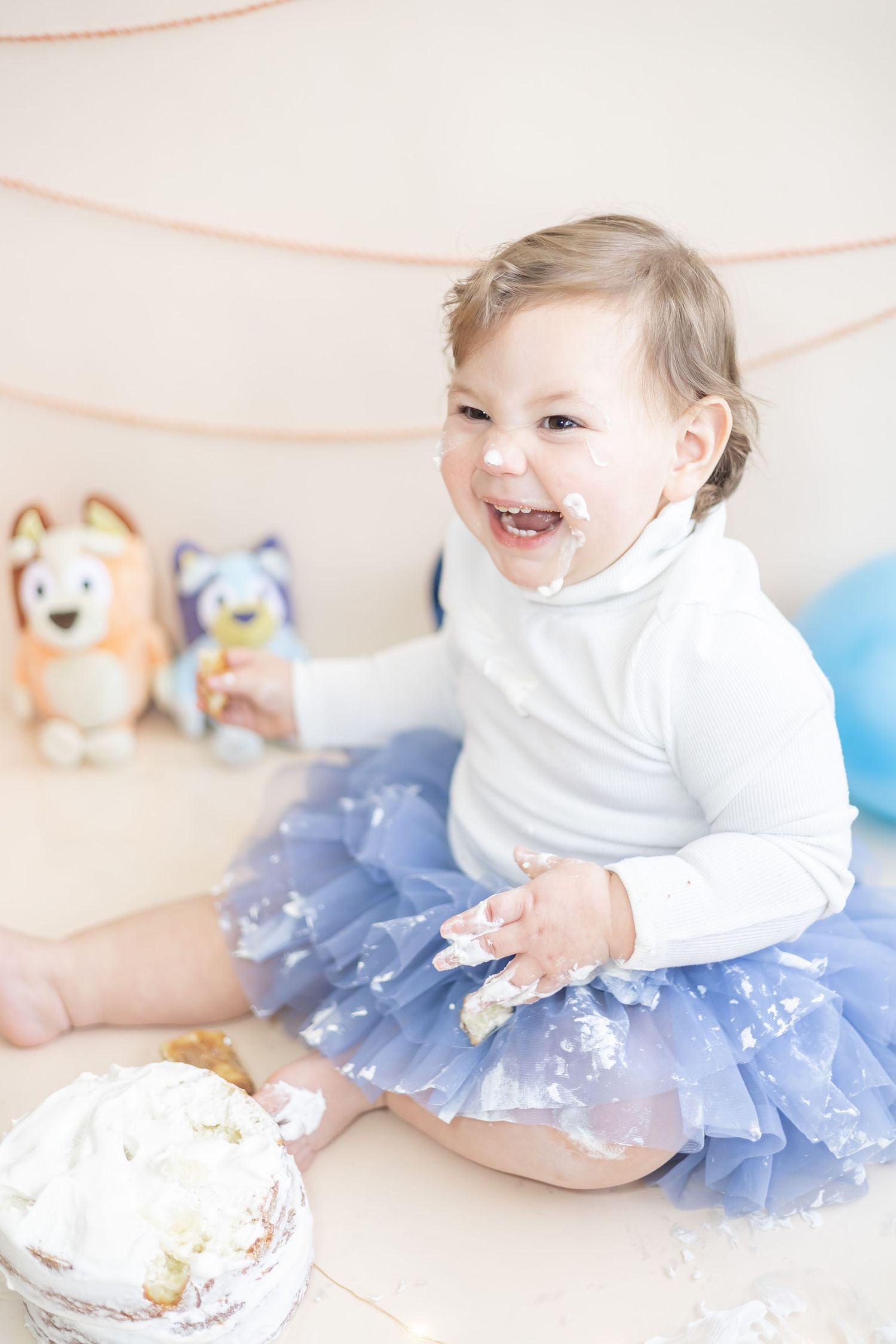 Baby in white shirt and blue tulle skirt sitting with frosted cake during first birthday cake smash photo session.
