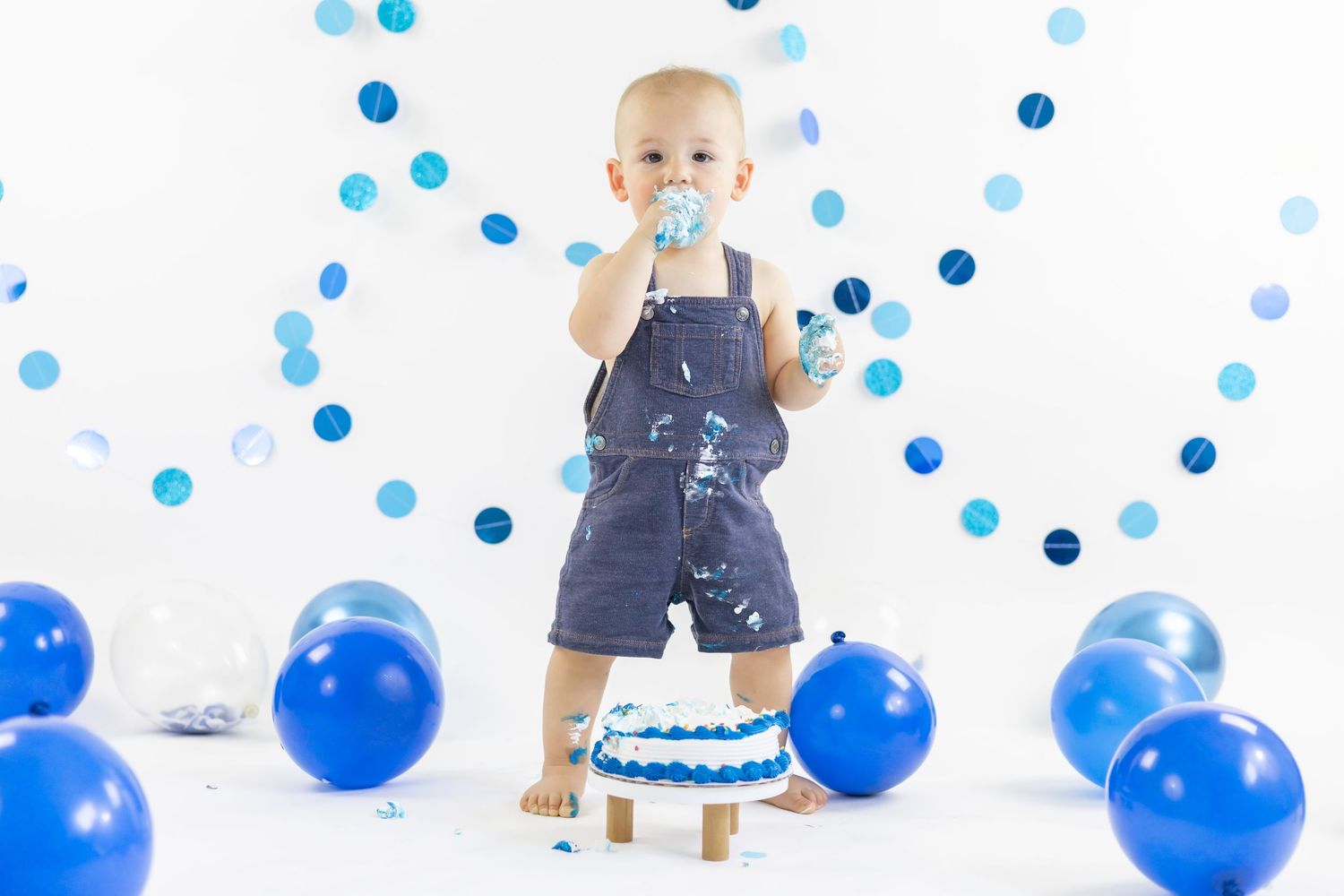 Baby in denim overalls celebrates first birthday with cake and blue balloons scattered on white floor.