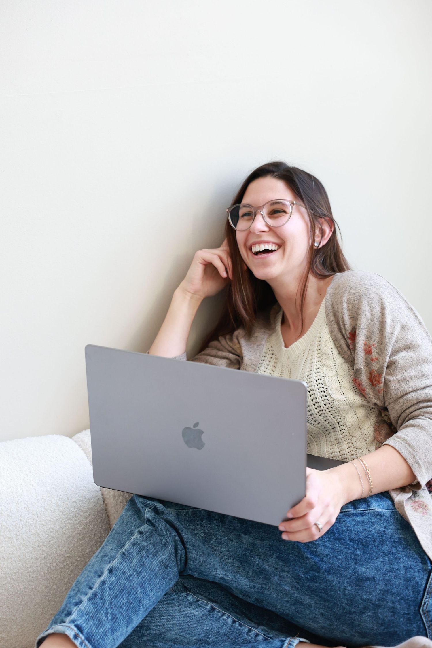 Person wearing glasses and a sweater sits casually with MacBook laptop against white wall, laughing while working.
