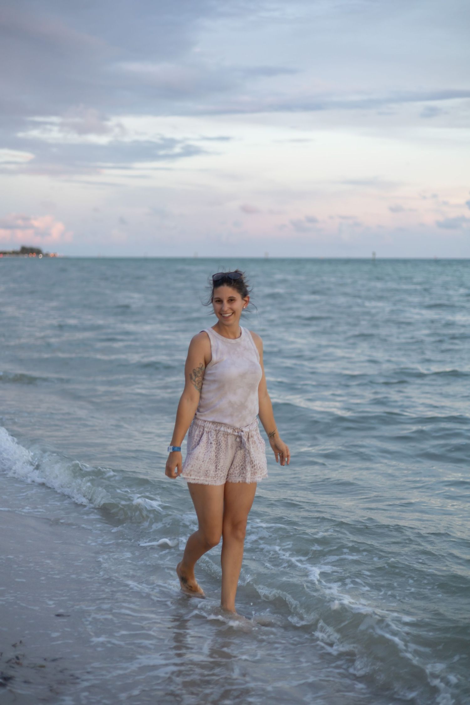 A person in light colored clothing wades into the shallow ocean water at dusk with a cloudy sky above.