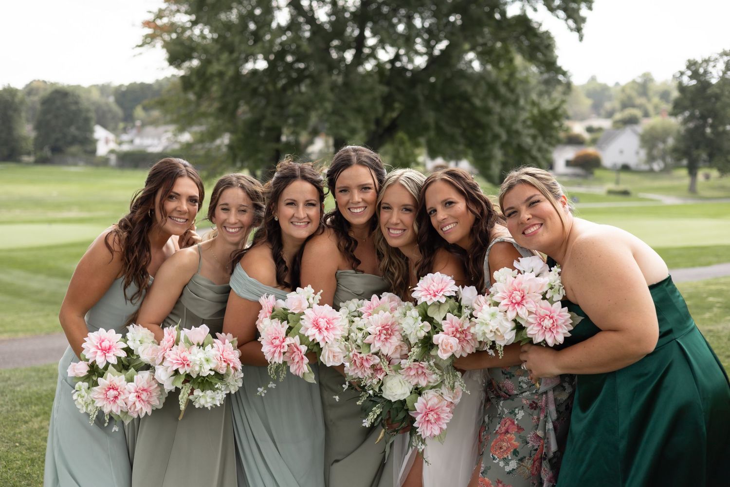 Bridesmaids in emerald green dresses pose together with pink floral bouquets at an outdoor wedding.