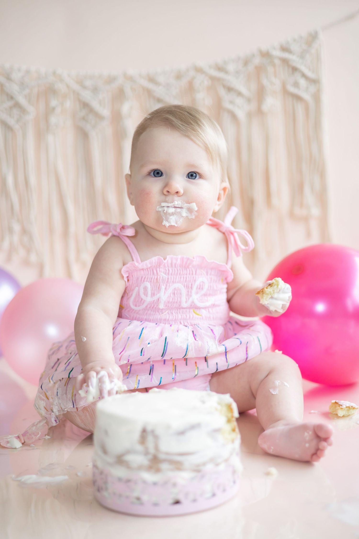 First birthday cake smash photography session with pink dress, balloons and white frosting decorations.