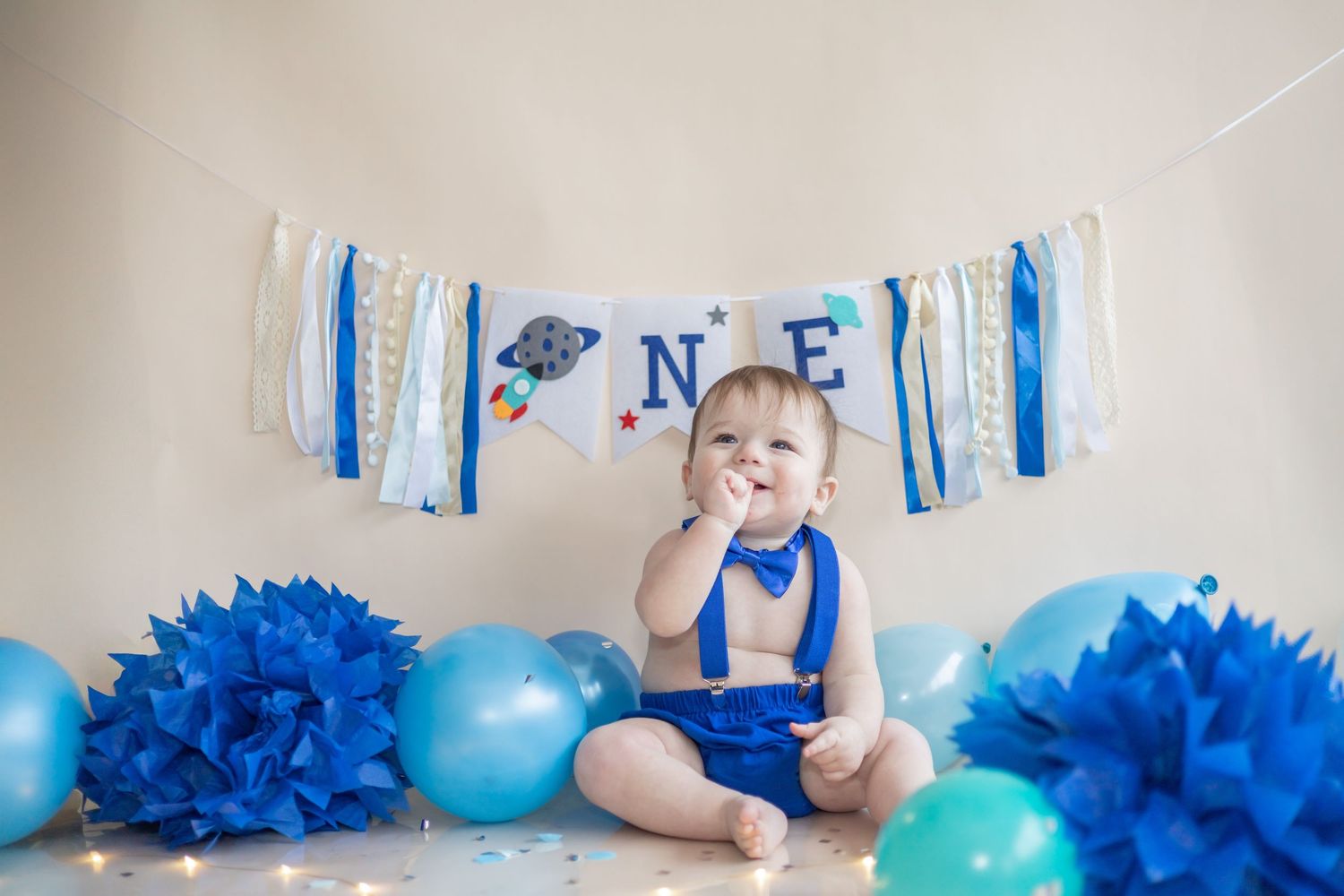 First birthday celebration setup with blue and white decorations including a banner, balloons and tissue paper pom poms.