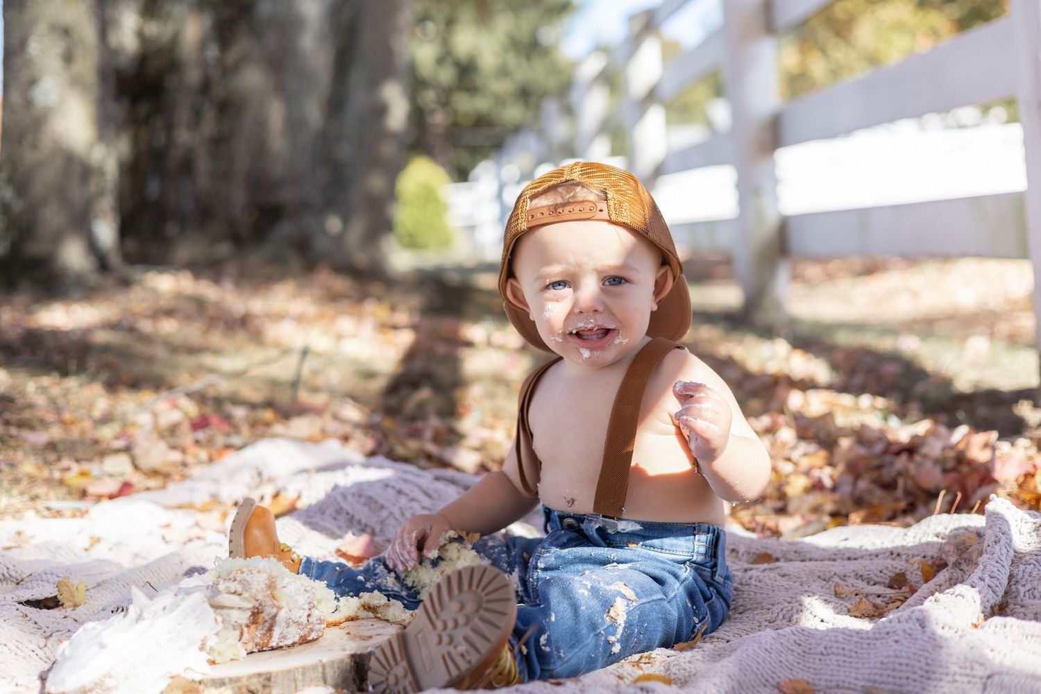 Small child wearing overalls and an orange cap sits on a blanket surrounded by autumn leaves near a white fence.