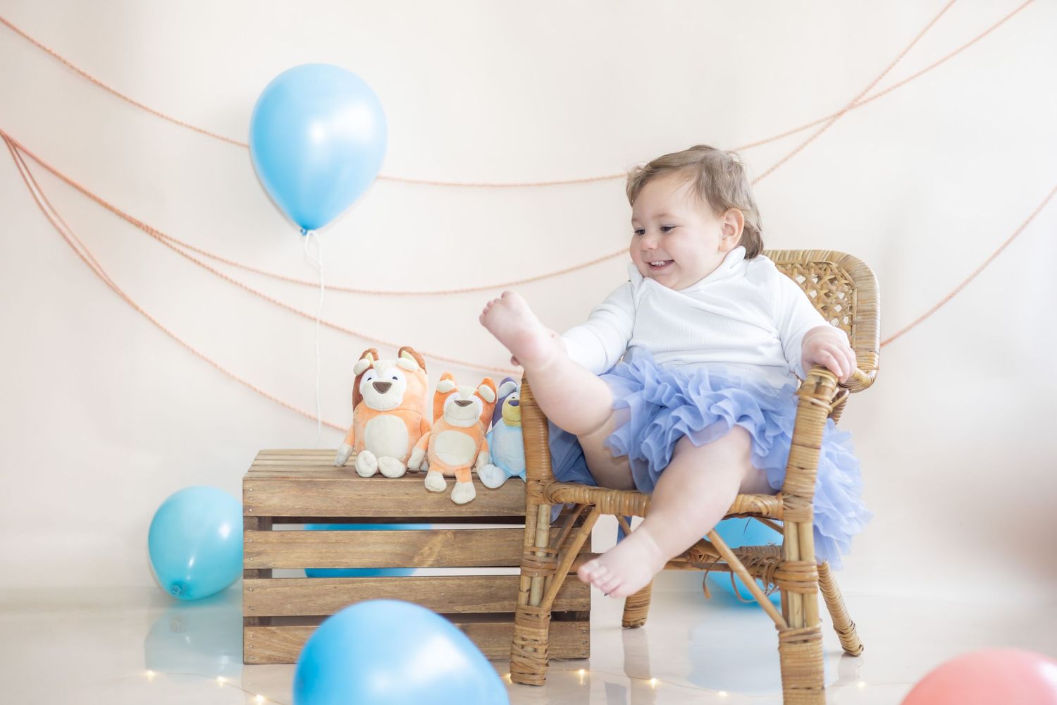 A baby sits in a wooden rocking chair surrounded by blue balloons and small stuffed animals on wooden crates.