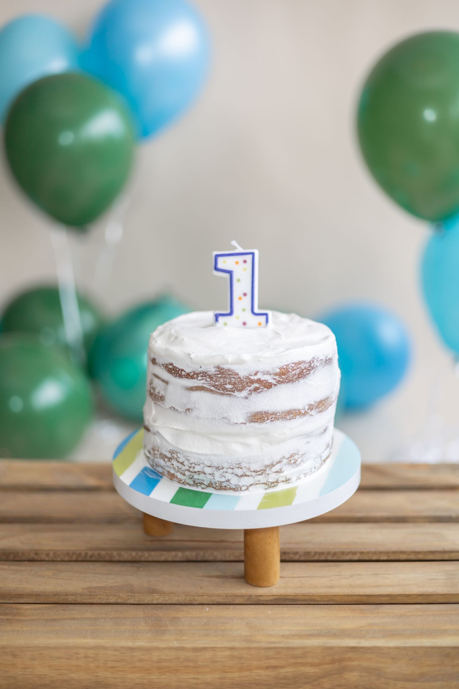 A rustic naked cake with a blue number one candle sits on a wooden cake stand with green and blue balloons in background.