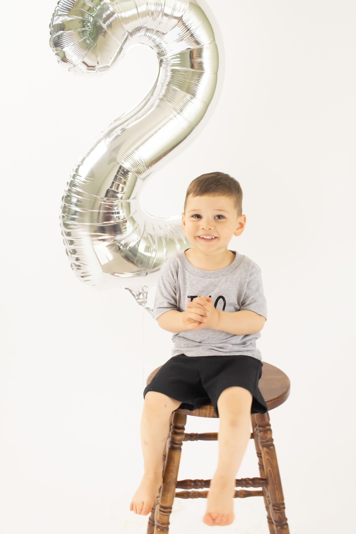 A toddler sits on a wooden stool with silver number 2 balloons in the background while wearing a gray t-shirt.
