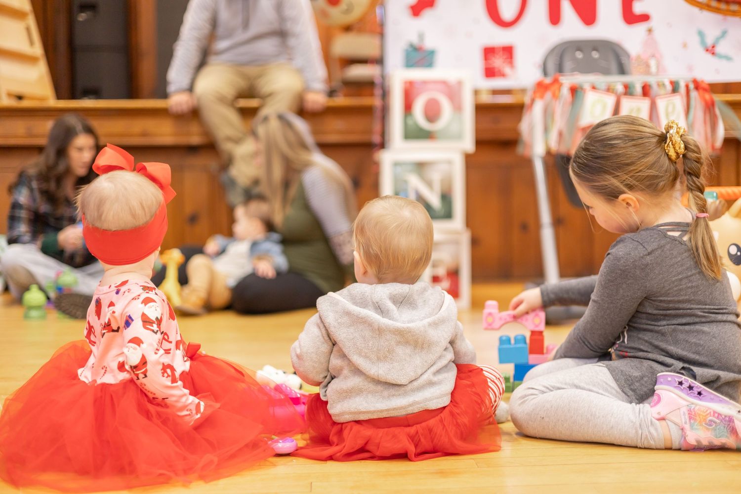 Young children sitting on the floor in a wood-paneled room with Christmas decorations at a holiday event.