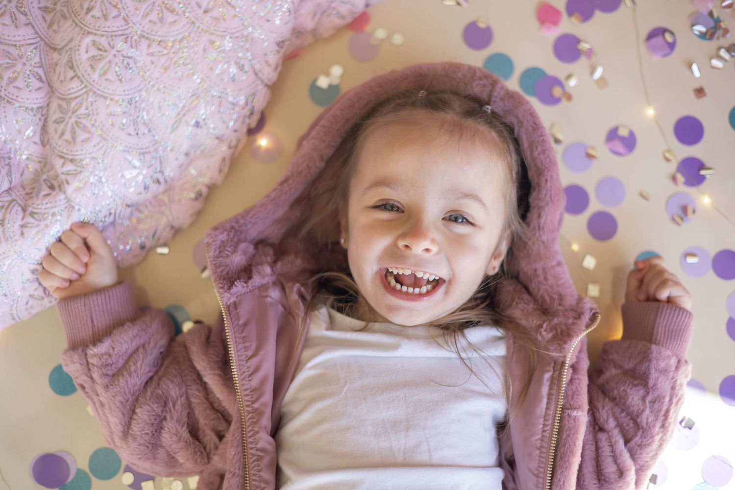 A child lies on the floor laughing joyfully surrounded by colorful confetti and wearing a cozy pink sweater.