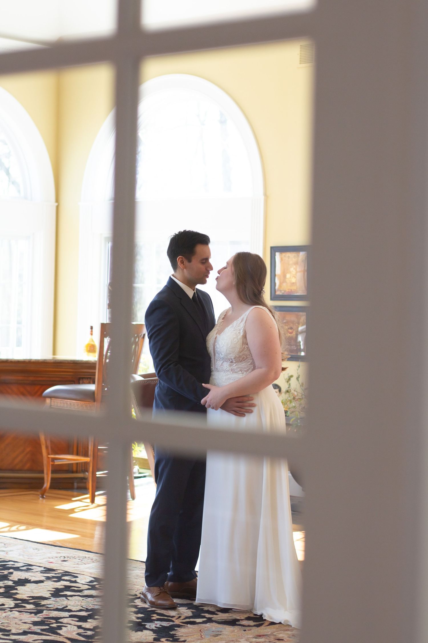 A couple in wedding attire share an intimate moment in an elegantly decorated room with arched windows and natural light.