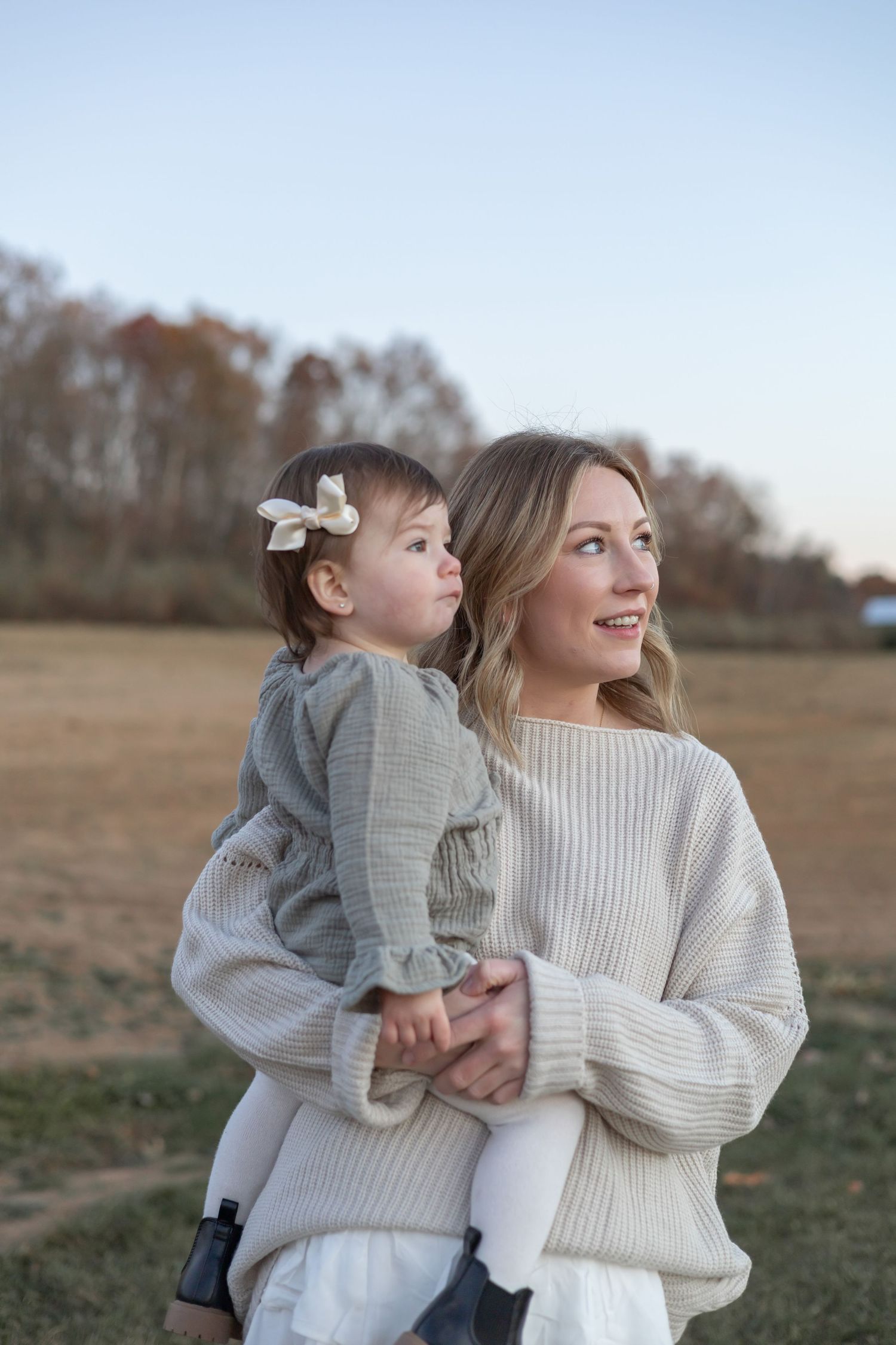A cozy autumn portrait of parent and child in warm sweaters standing together in a golden field at sunset.
