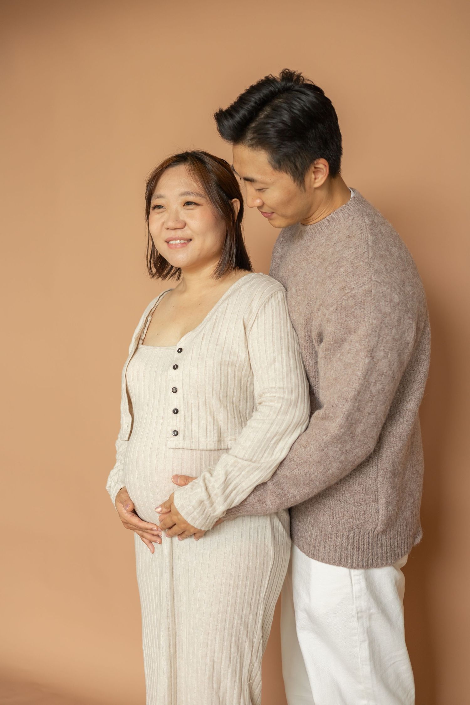 A couple in casual attire shares an intimate moment against a brown backdrop as they expect a baby.