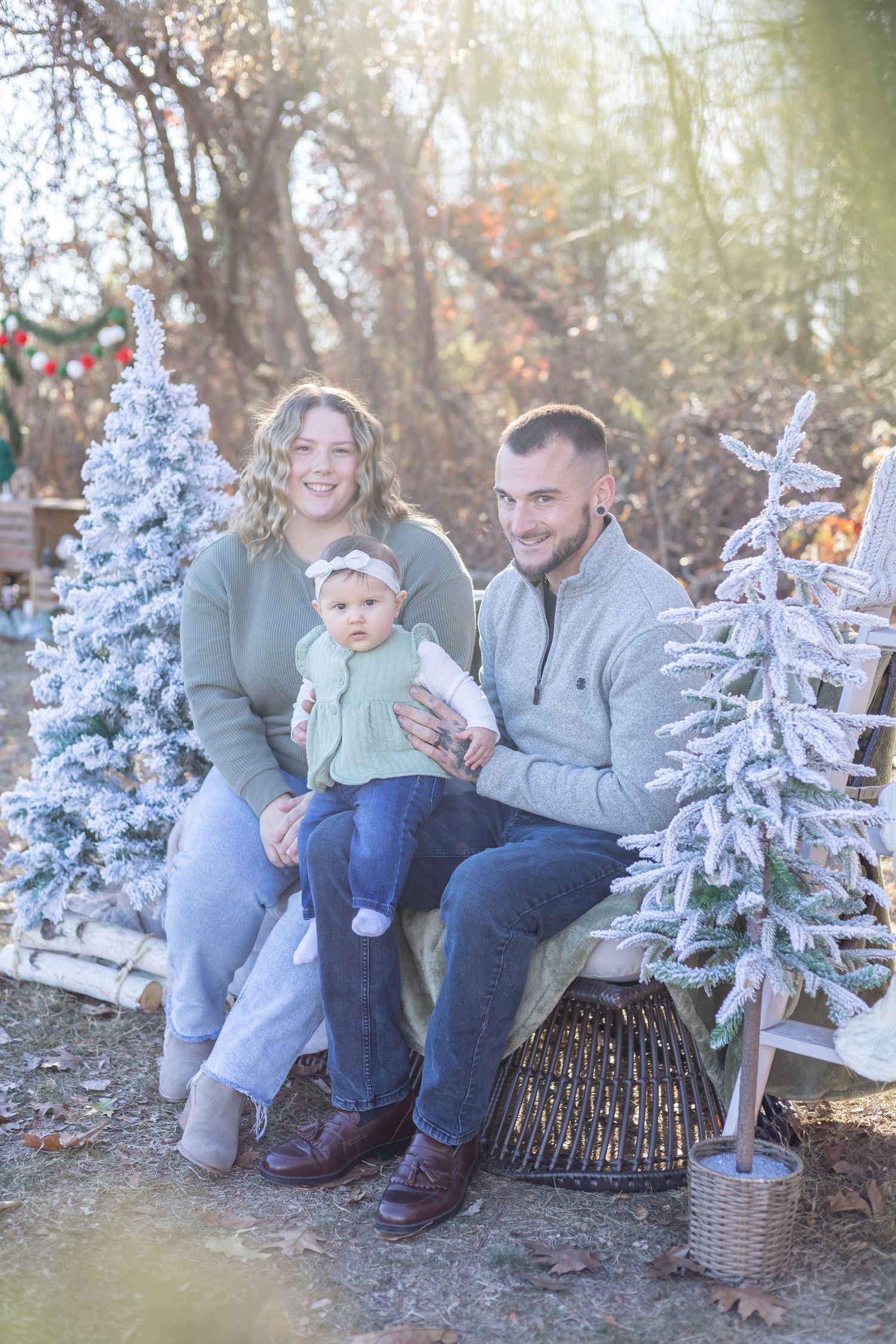 A family sits together on a rustic log bench surrounded by snow-dusted evergreen trees in a winter outdoor setting.