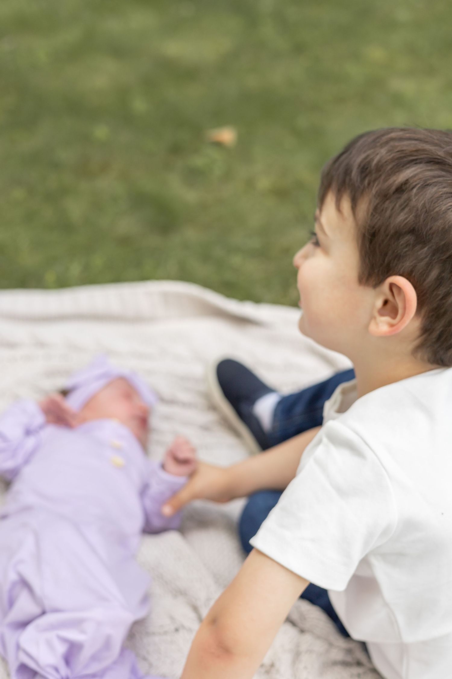 Child in white shirt lies on blanket next to baby in purple clothing while relaxing outdoors on grass.