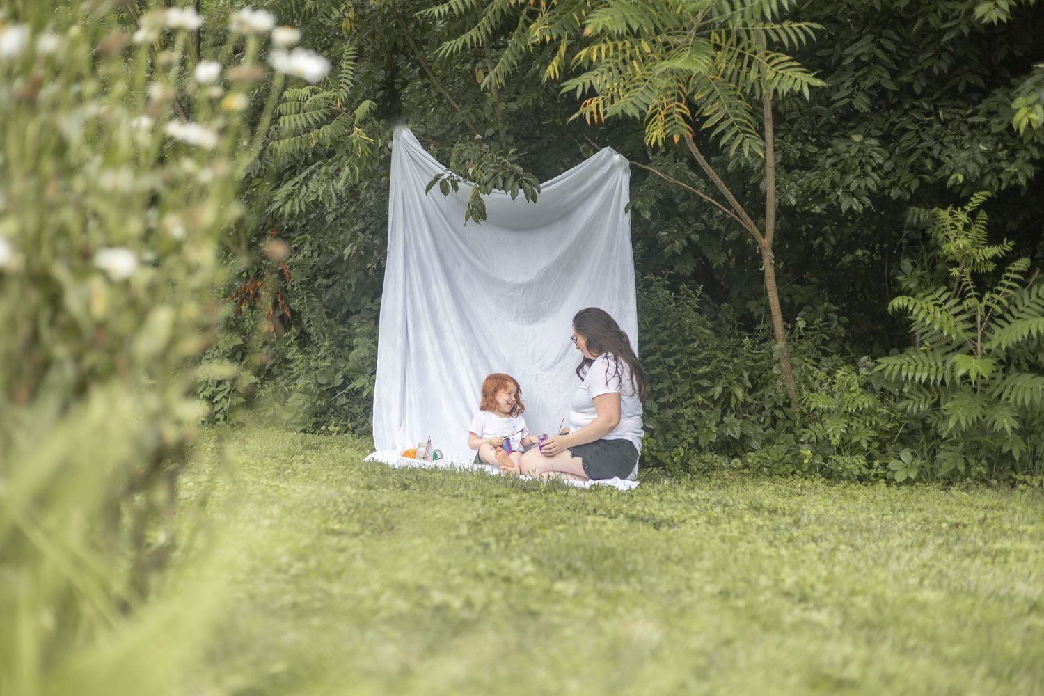 A group of images hanging on white cloths strung between trees in a lush green garden setting.
