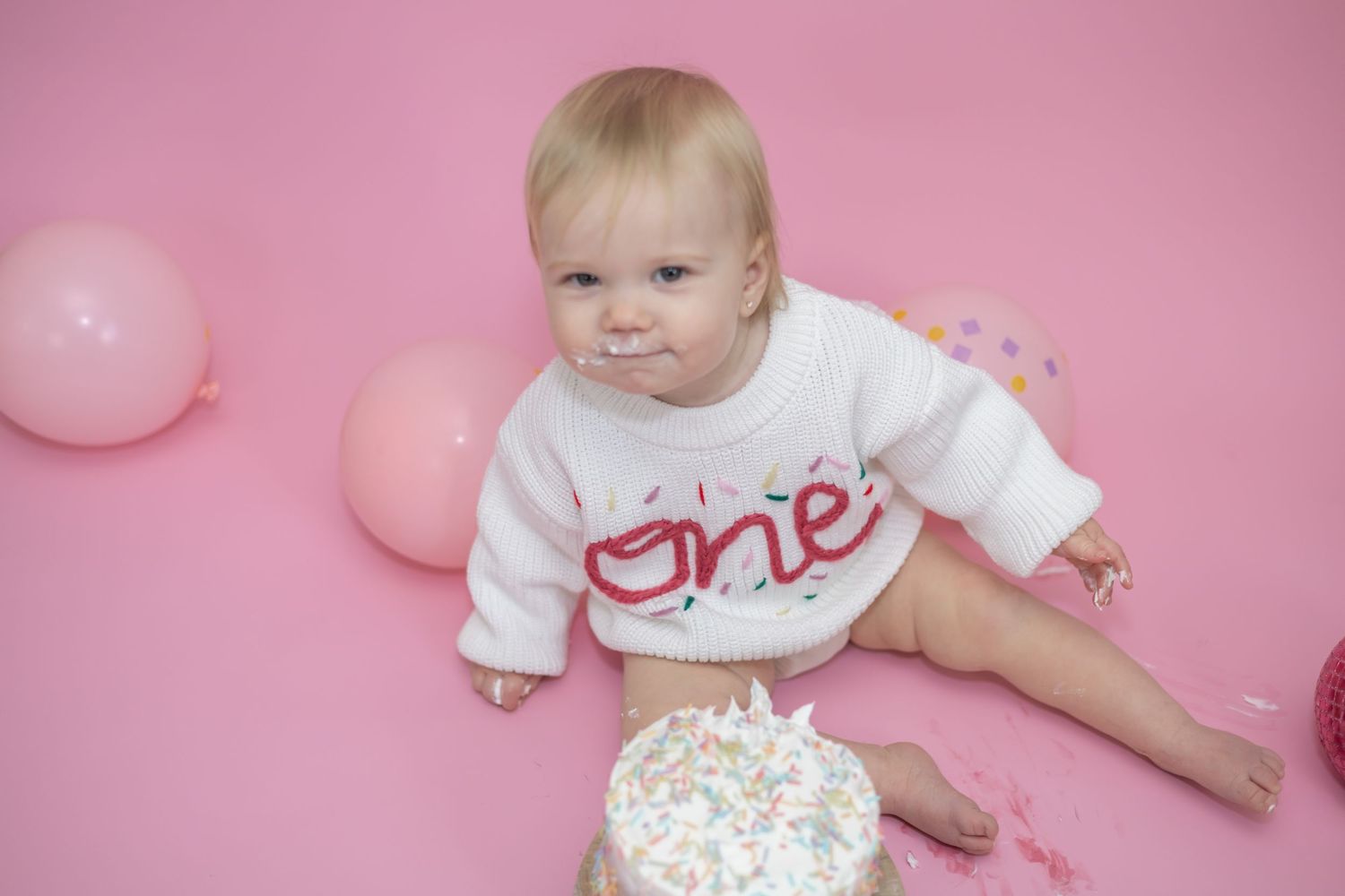 A baby in a white 'ONE' shirt sits among pink and white balloons during a first birthday photoshoot.
