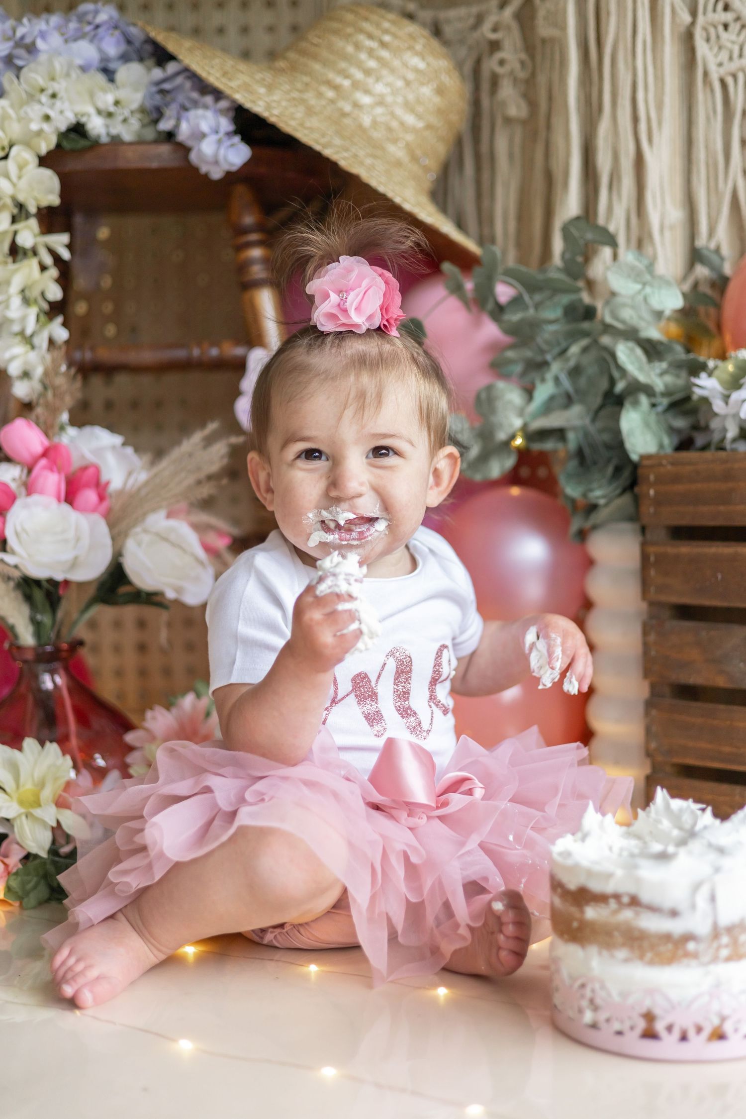 A small child in a pink tutu sits on the floor with a smash cake and decorative floral arrangements for a special celebration.