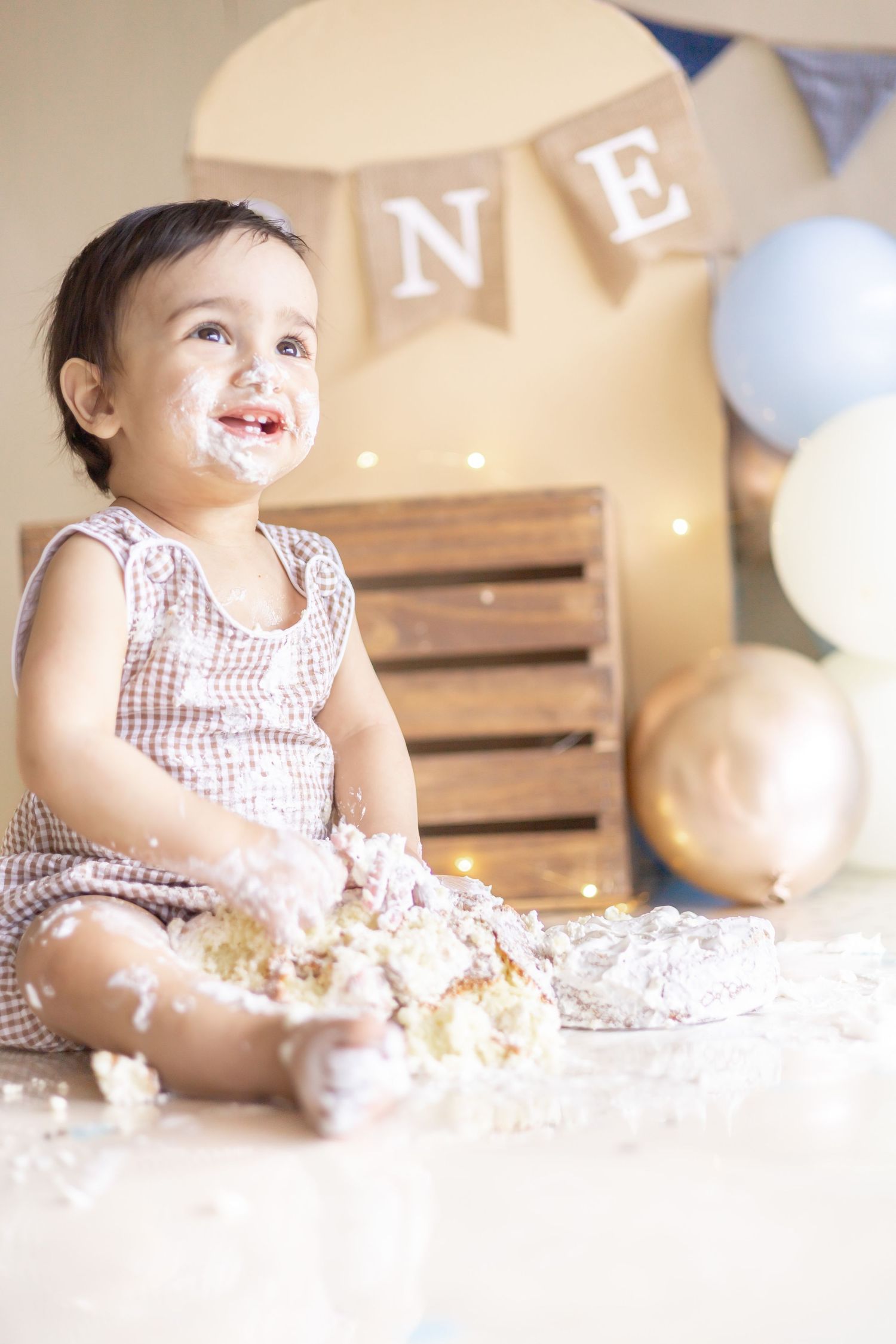 Joyful toddler in checkered dress plays with cake and flour during first birthday photoshoot with party decorations.
