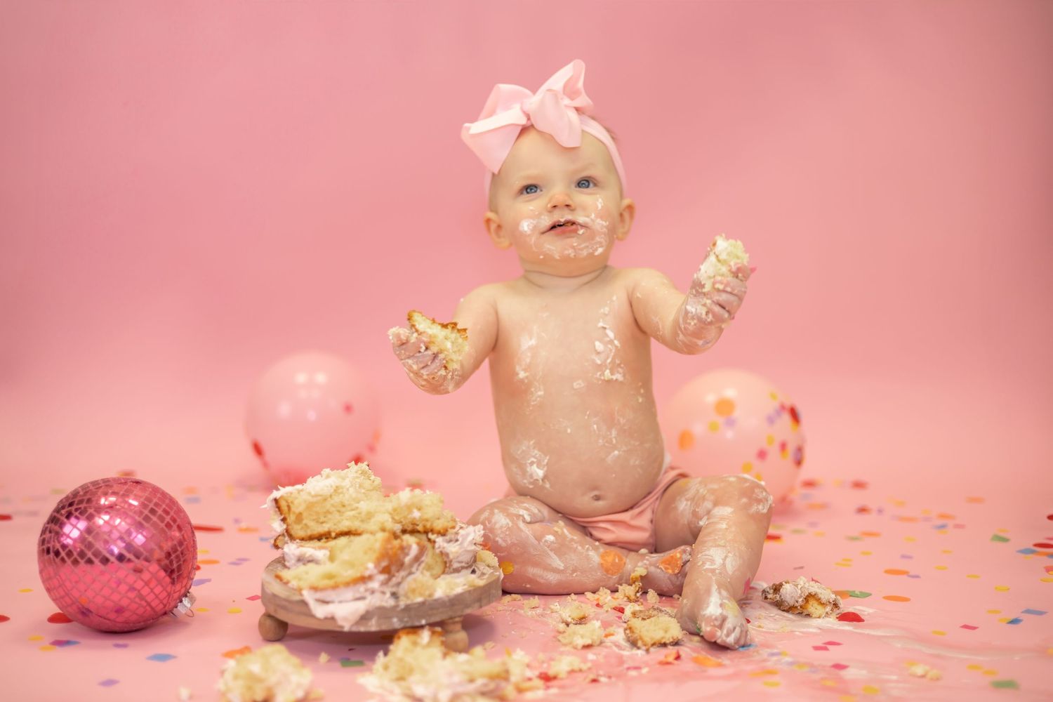 Messy cake smash photo with glitter-covered baby, destroyed cake, pink disco ball and confetti on pink background.